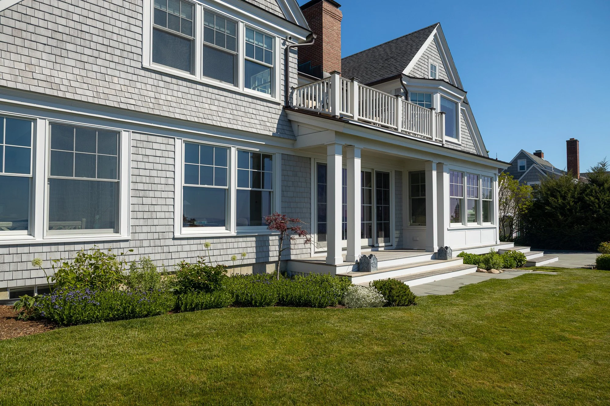 A large house with a sloped roof, multiple windows, a small balcony, and a porch with steps, surrounded by a green lawn and garden plants under a clear blue sky.
