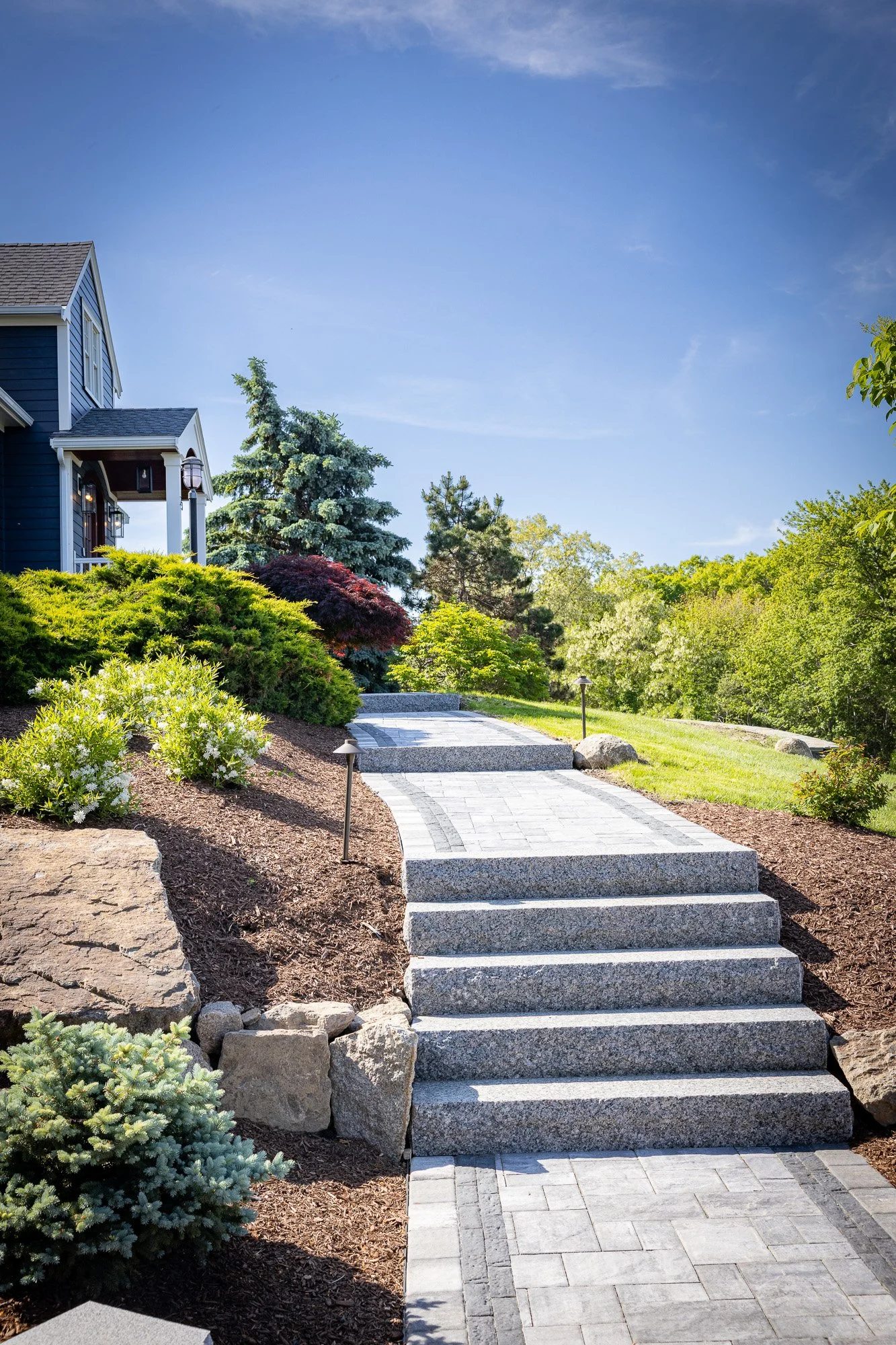 A landscaped front yard with a staircase leading up from the walkway, surrounded by bushes, trees, and a house with blue siding and white trim in the background, under a blue sky.