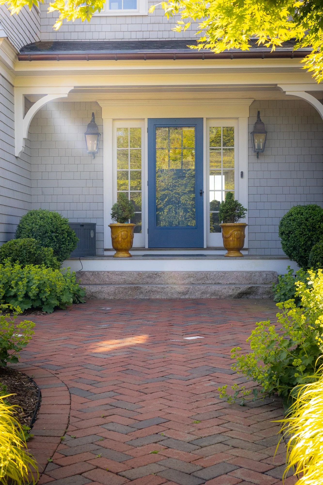Front porch of a house with a blue door, side windows, hanging lantern lights, potted small trees, surrounded by green bushes, and a curved brick walkway leading to the porch.