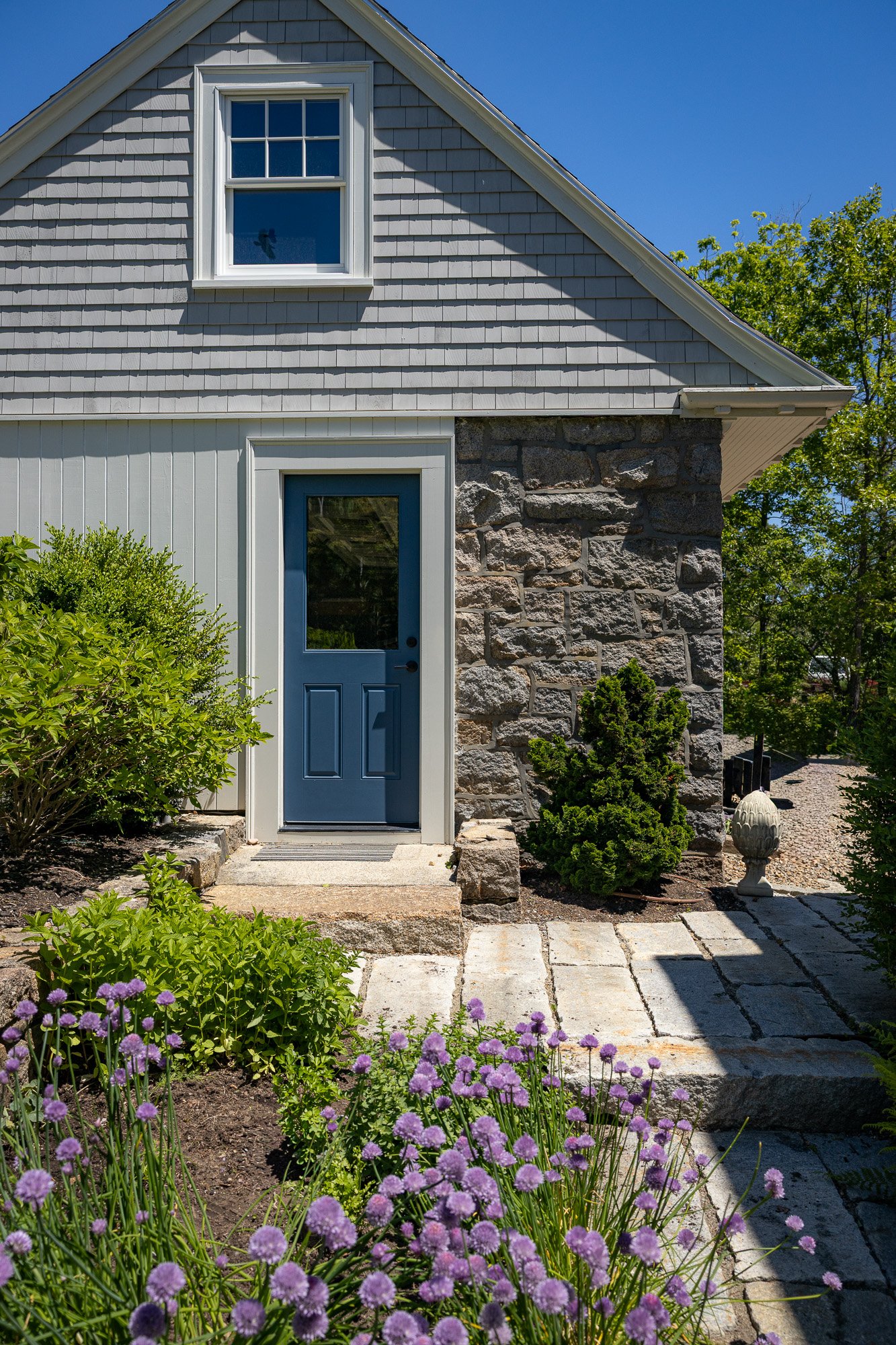 Front door of a house with grey shingles, a stone wall, and a blue door, surrounded by green bushes and purple flowers under a clear blue sky.
