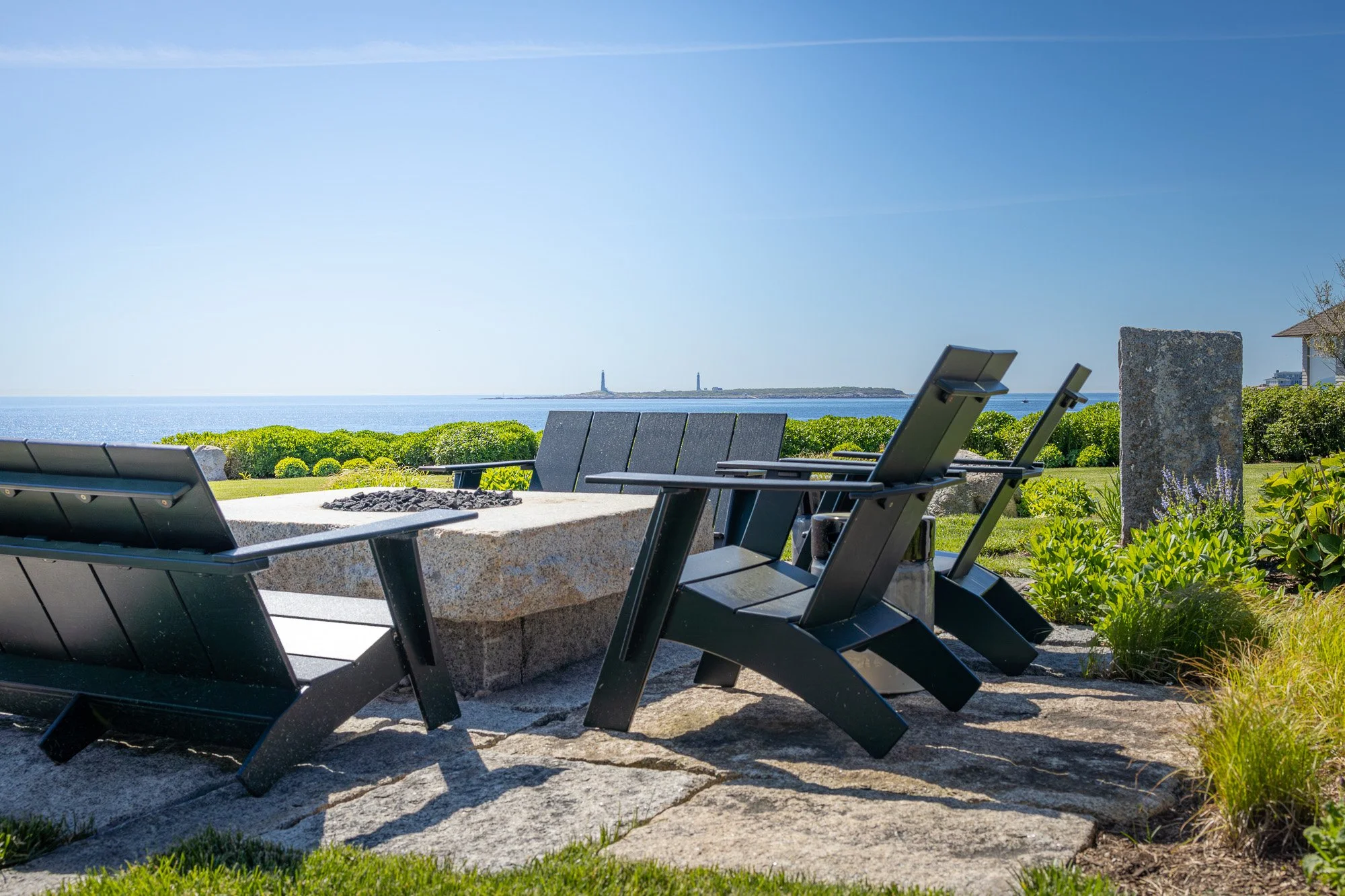 Outdoor seating area with metal chairs around a fire pit, overlooking a body of water with a lighthouse in the distance under a clear sky.