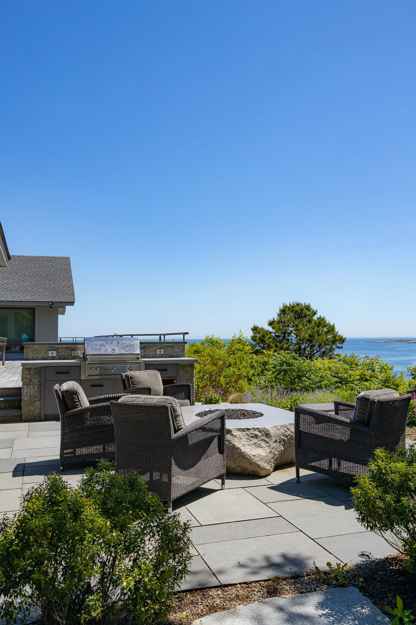 Outdoor patio with wicker chairs, a built-in grill, a stone fire pit, lush greenery, and a scenic ocean view with a clear blue sky.