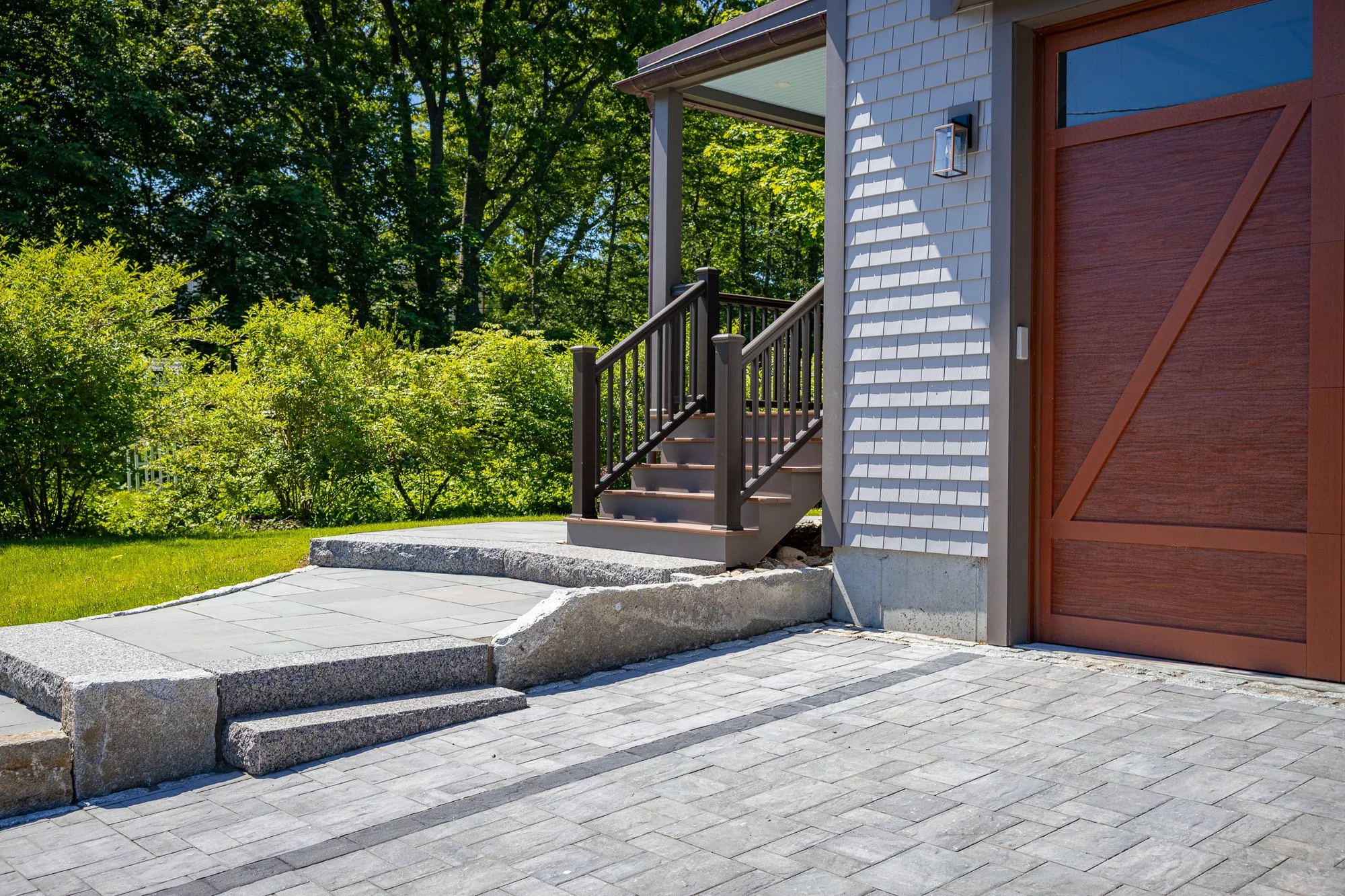 A modern house exterior featuring a raised porch with steps, a brown wooden garage door, a capsule light fixture, and a lush green tree background.