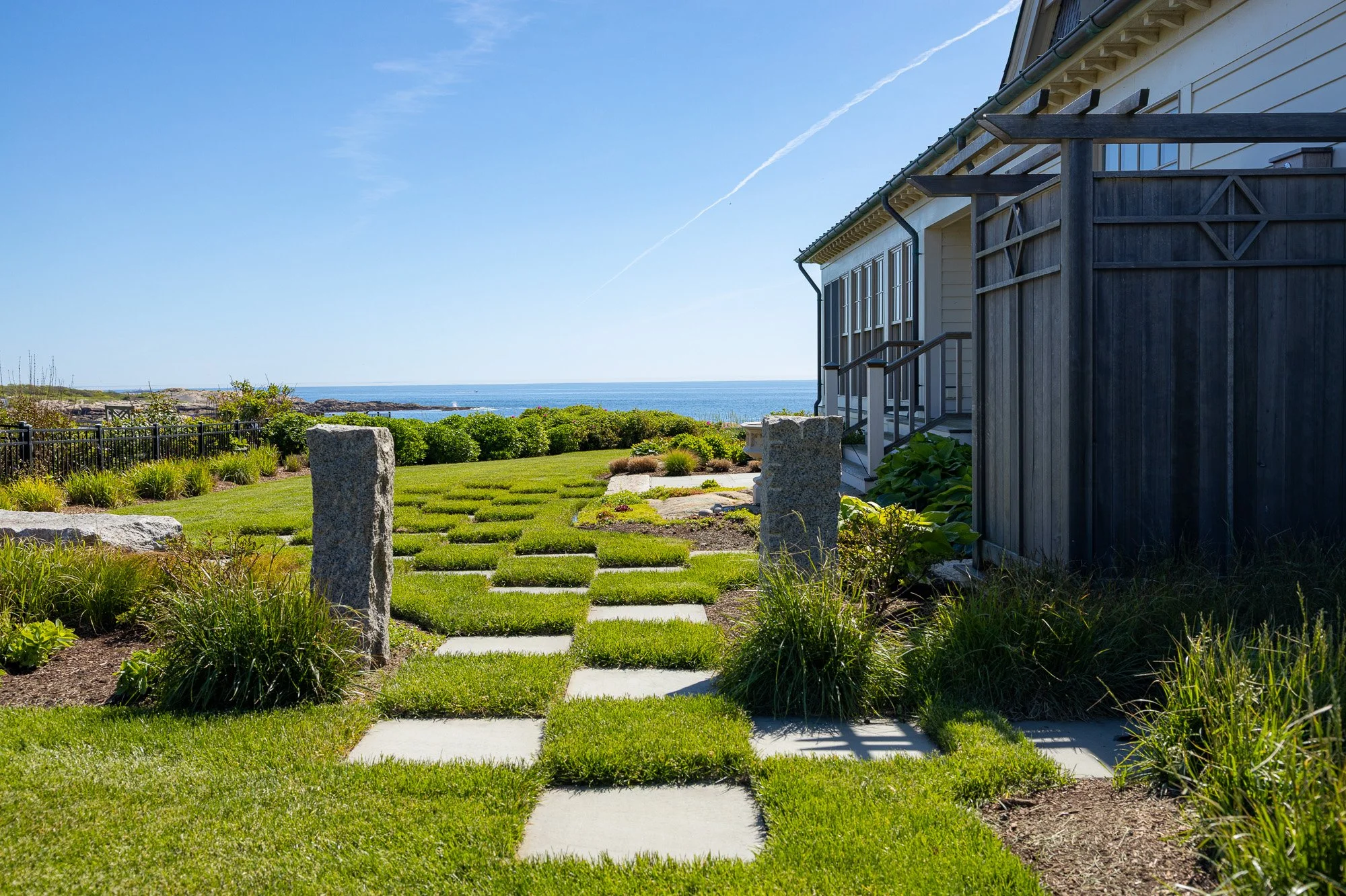 A house with a wooden fence and a garden leading to the ocean in the distance. The sky is clear and blue.