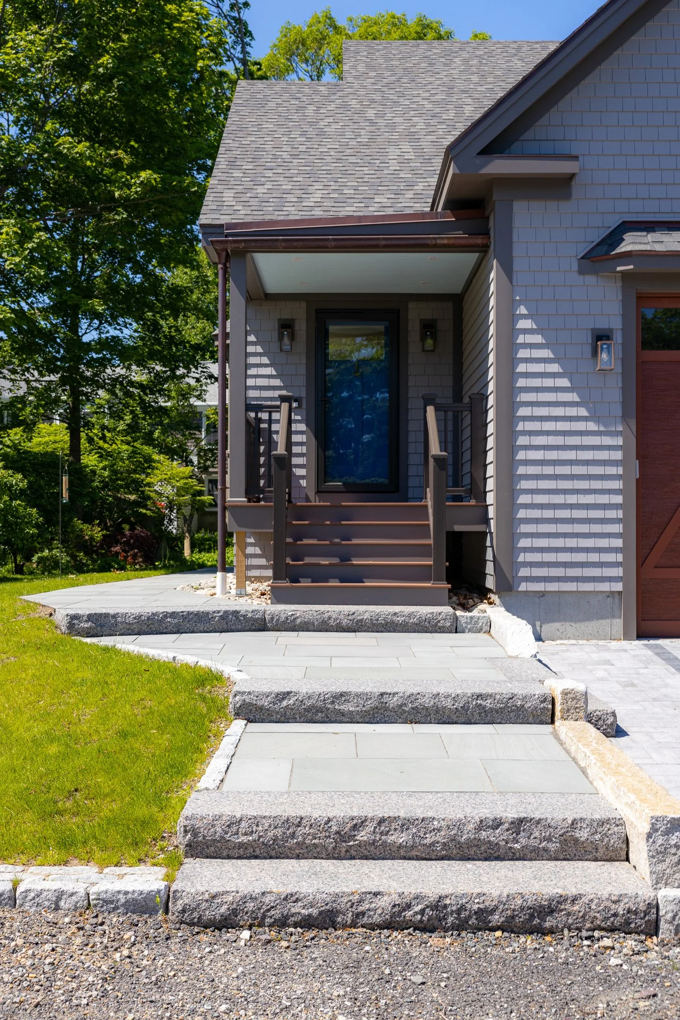 Front view of a house with grey shingles, a small porch with steps, and a pathway leading to the entrance. Green trees and a clear blue sky in the background.