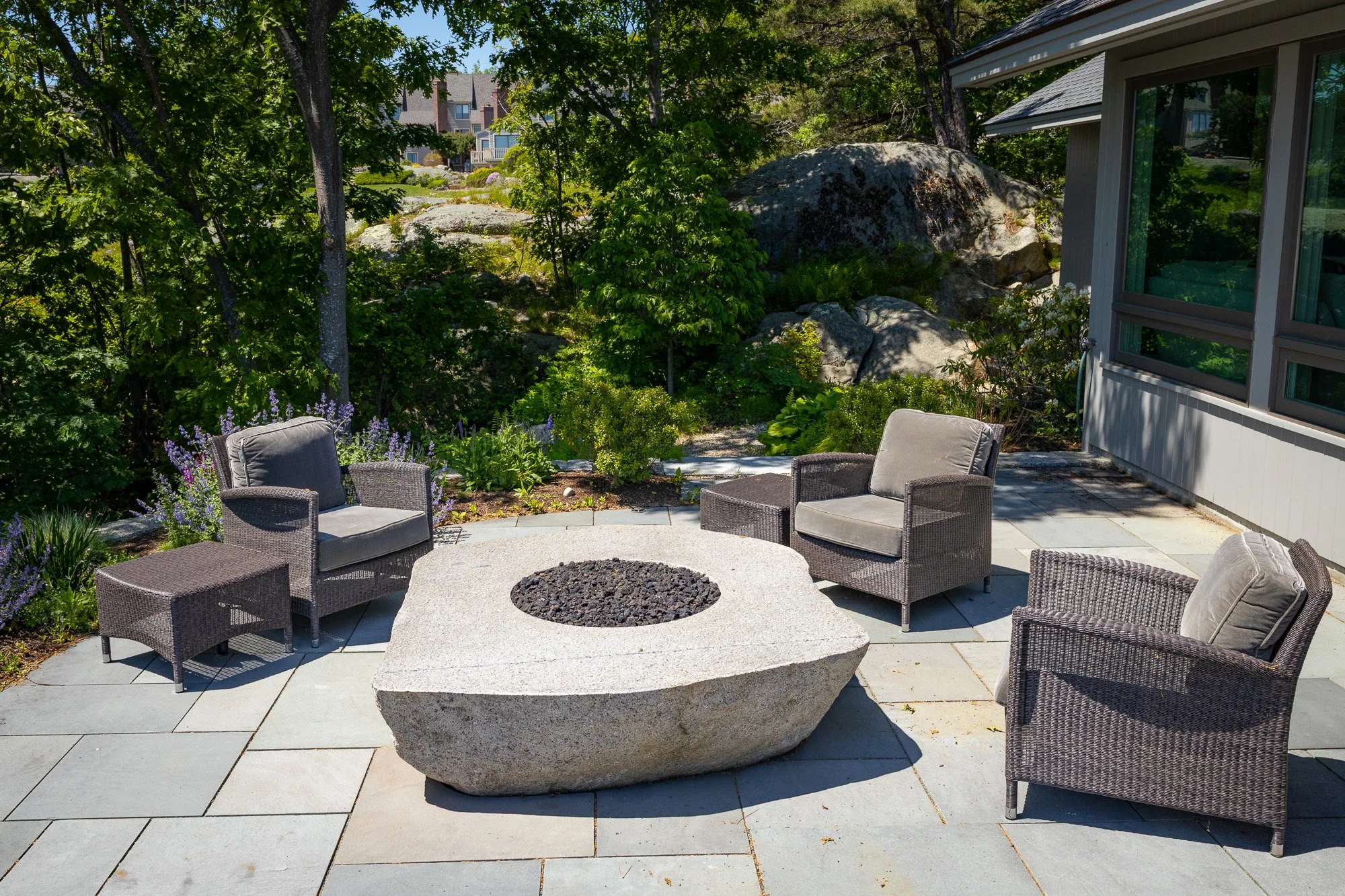 Outdoor patio with a circular fire pit, surrounded by wicker chairs with gray cushions, lush green trees, and a house with large window on the right. Large rocks and a garden are visible in the background.