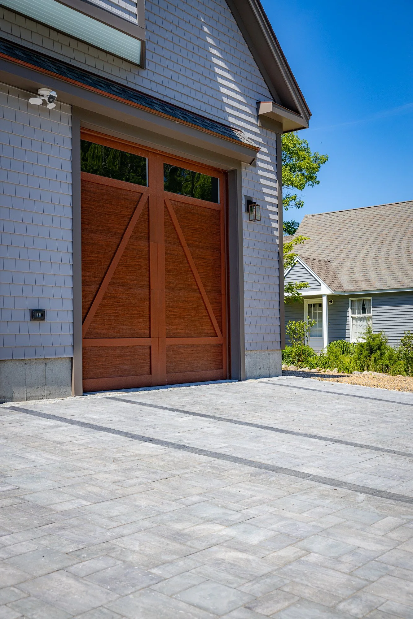 The image shows the front of a modern house with a garage door made of wood and glass, a security camera mounted on the wall, and a paved driveway leading up to the garage. There are other houses and trees visible in the background under a clear blue