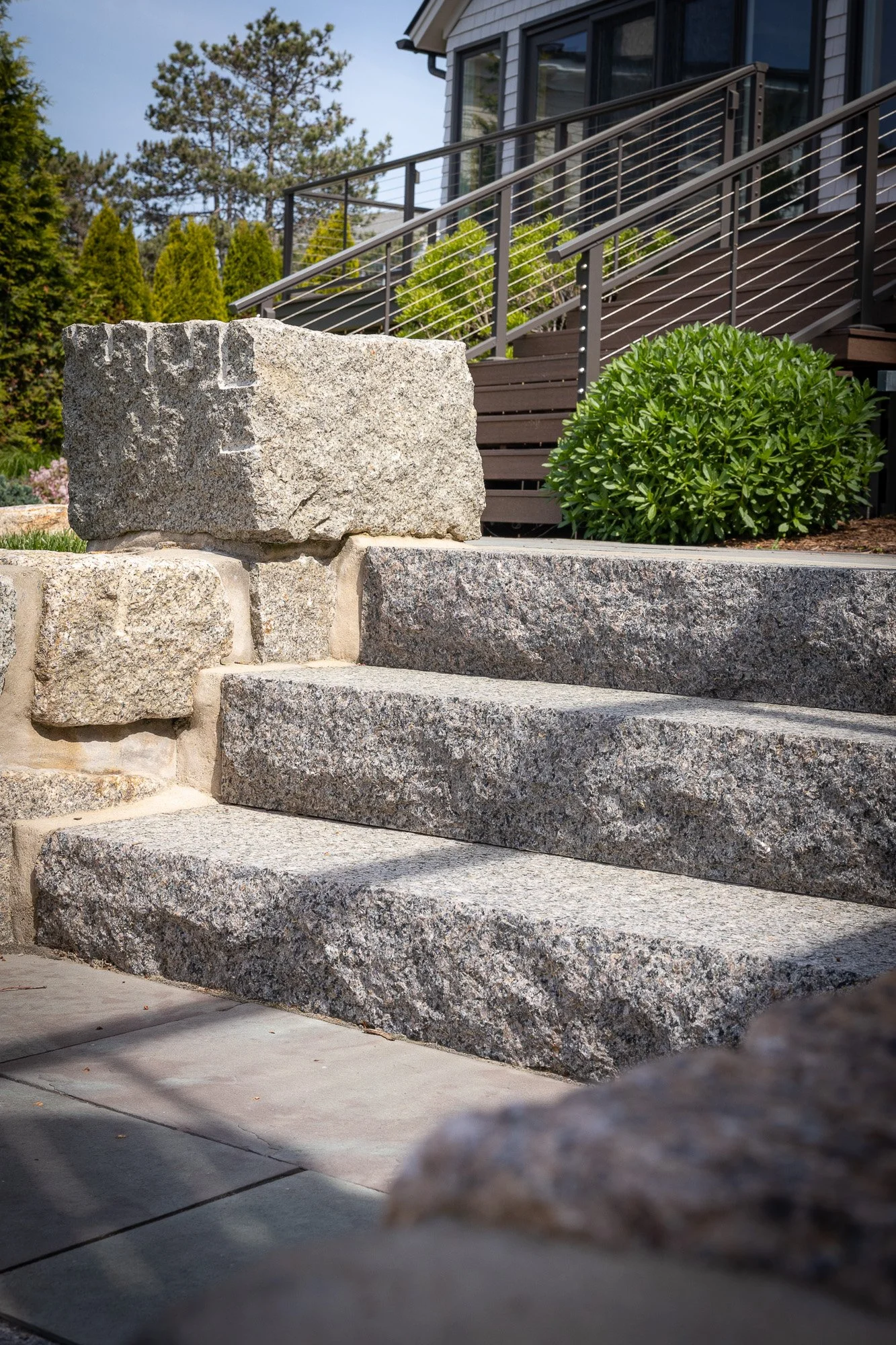 Stone staircase leading to a house with a metal railing, surrounded by greenery and trees in the background.