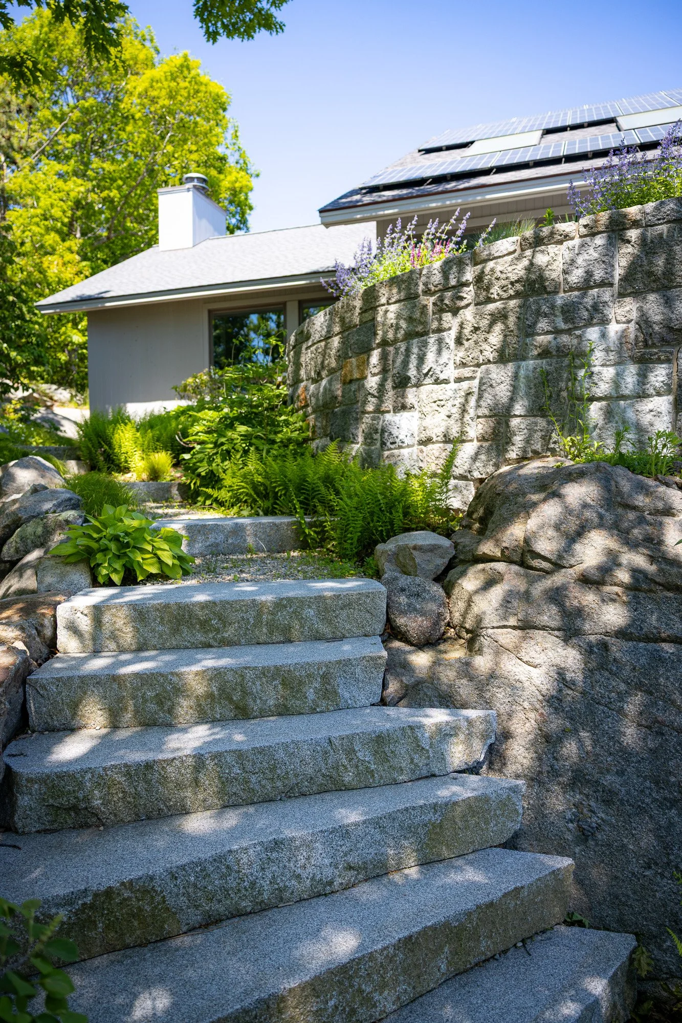 Stone steps leading up to a house with a garden, greenery, and a stone wall. The house has solar panels on the roof and a chimney. Trees and plants are around the steps, with a clear blue sky overhead.