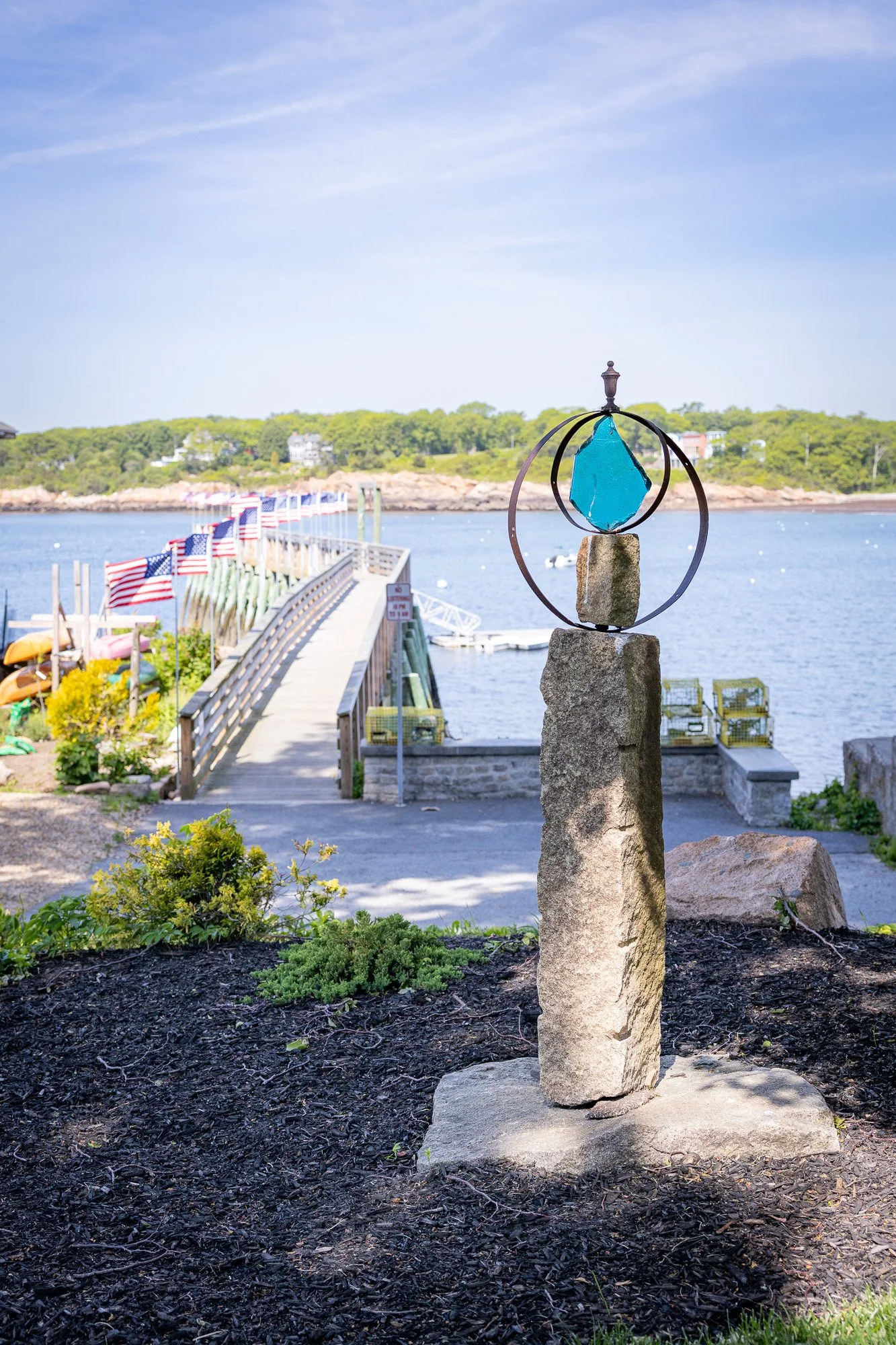 Metal sculpture with blue glass, concrete, and metal rings on top of a stone pillar by the water with a dock, American flags, boats, and green hills in the background on a sunny day.