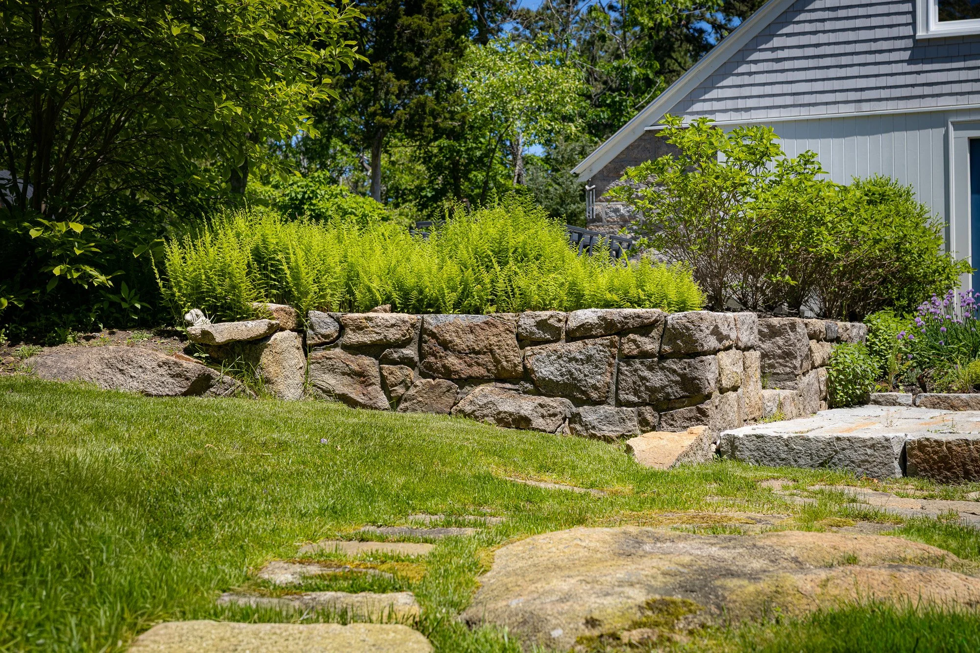 A landscaped front yard with a low stone retaining wall. Green bushes and small trees grow behind the wall, with a house in the background. The house has gray siding and a sloped roof. The yard has a well-maintained lawn with stepping stones leading 