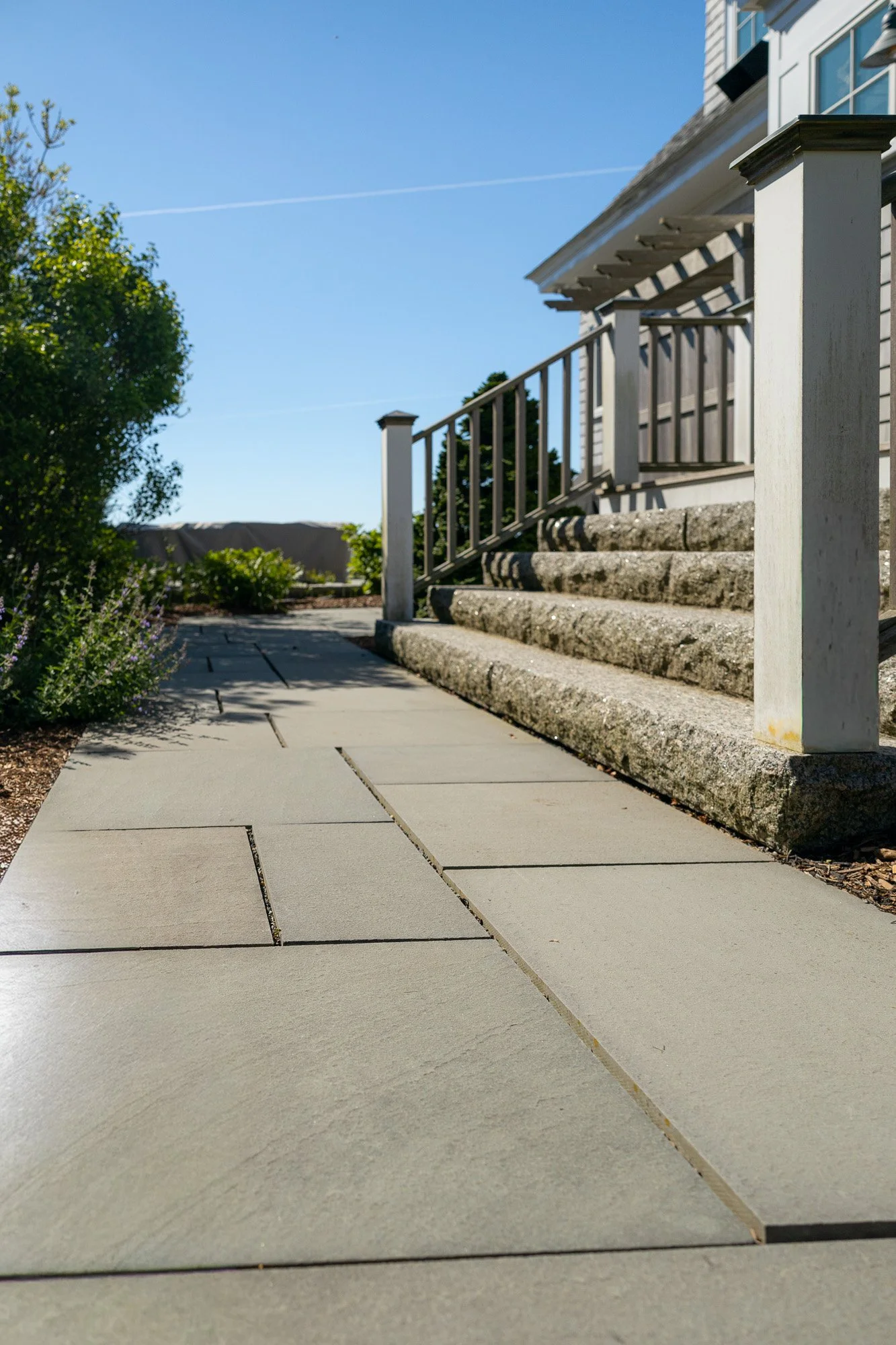 A front yard with concrete steps leading up to a house porch, surrounded by plants and trees, under a clear blue sky.