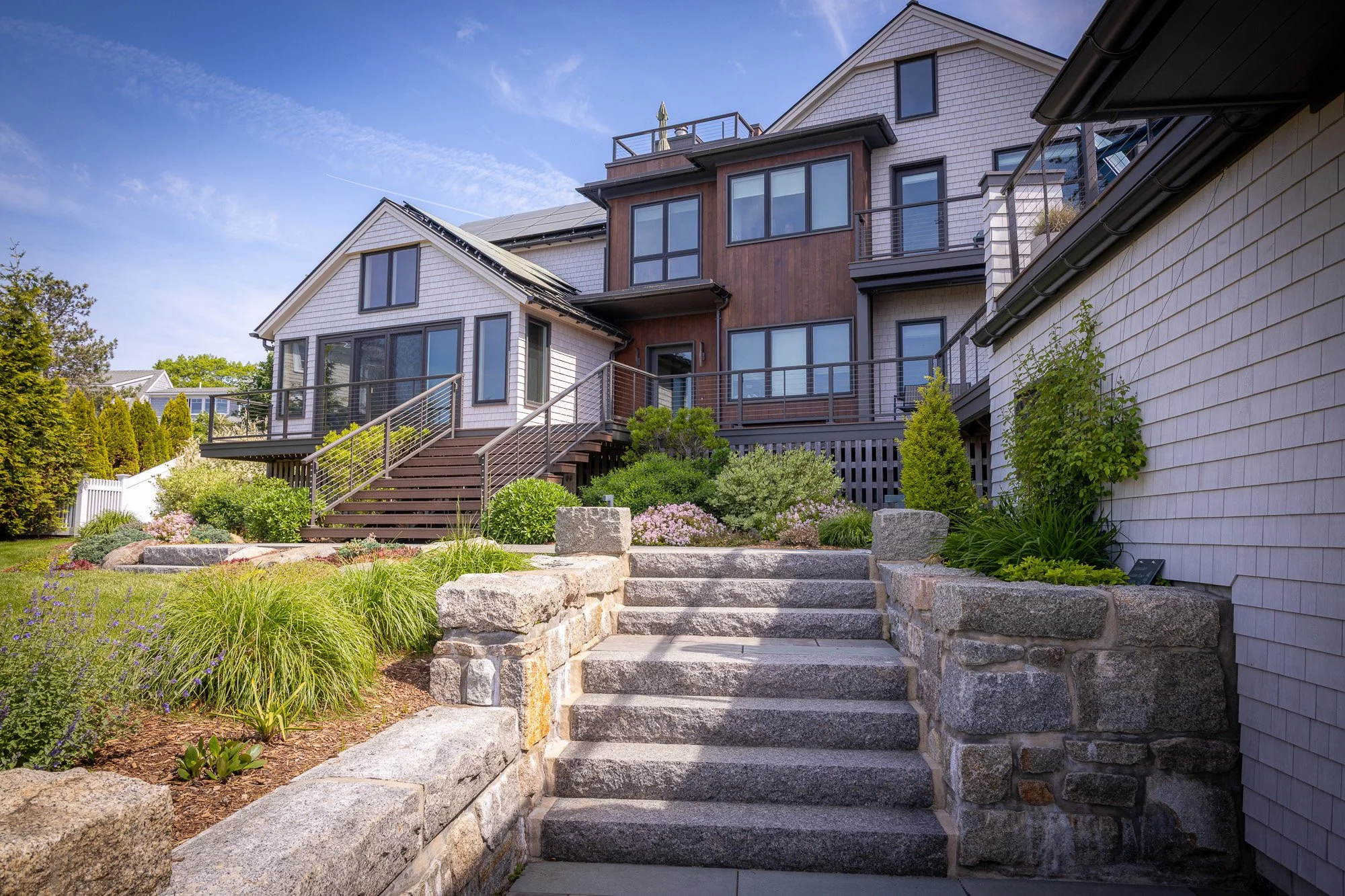 Backyard of a modern multi-story house with a landscaped garden, stone pathway, and stairs leading to a deck, under a blue sky with some clouds.