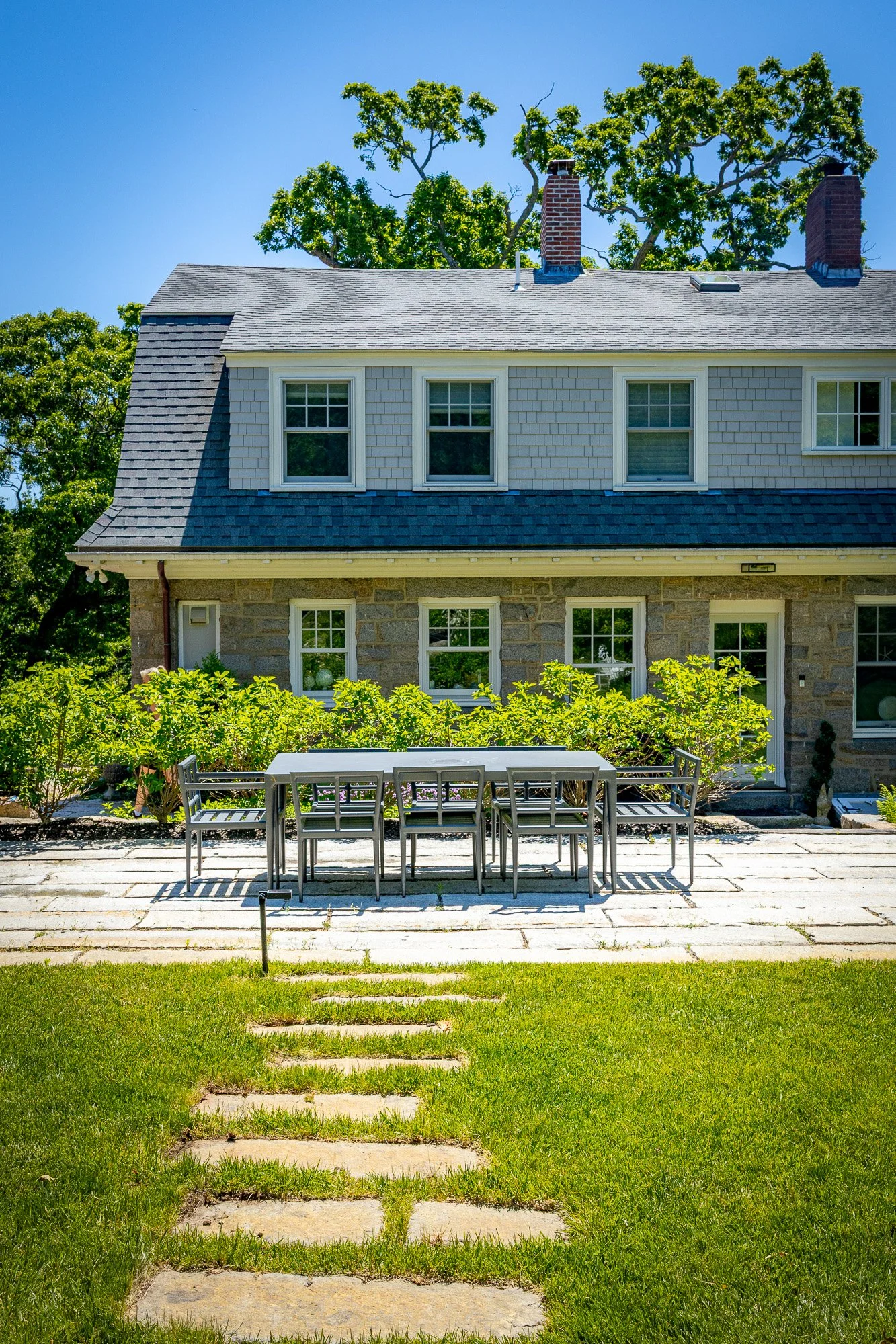 Backyard scene with green lawn, stepping stones, outdoor dining table and chairs, garden with bushes, house with gray roof and white and brick exterior, trees and chimney in the background under blue sky.
