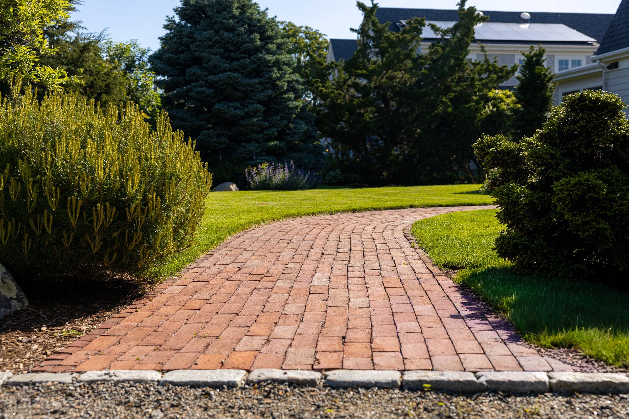 A brick pathway curves through a well-maintained front yard with green grass, bushes, and trees, with a house in the background.