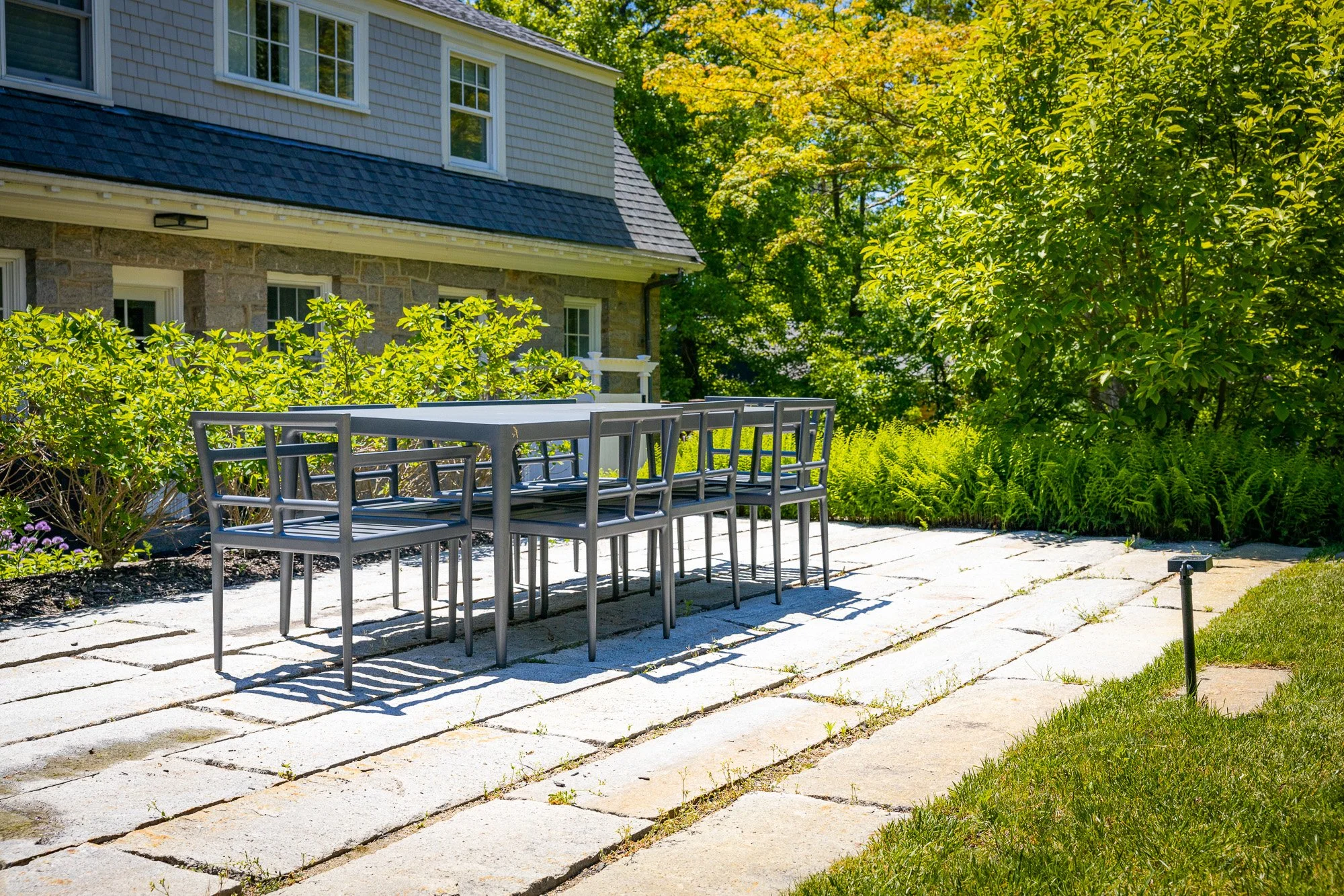 A patio with outdoor table and chairs, stone paving, surrounded by lush green plants and trees, with a house in the background.