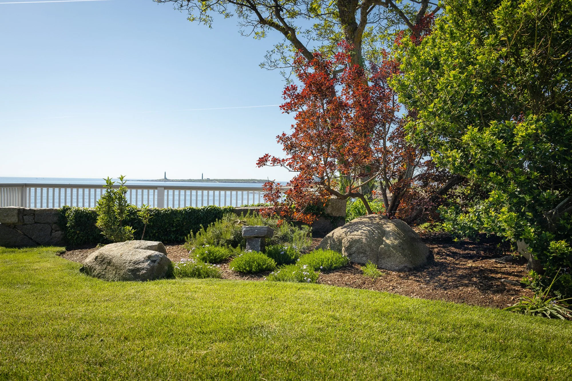 A landscaped garden with green grass, trees with green and red leaves, large rocks, and a view of water and a distant lighthouse through a white fence.