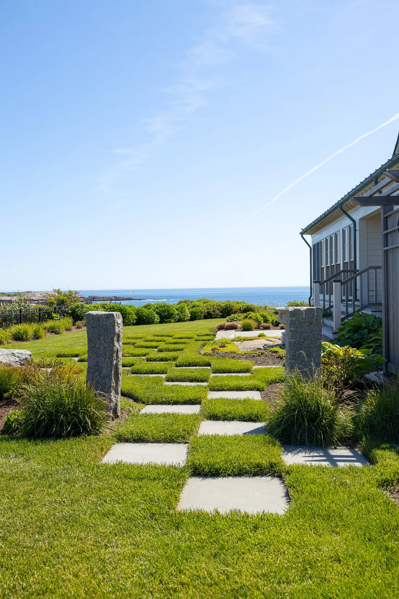 A garden with a stone pathway leading to the beach, surrounded by green grass, shrubs, and plants, with a house on the right, and the ocean in the background under a clear blue sky.