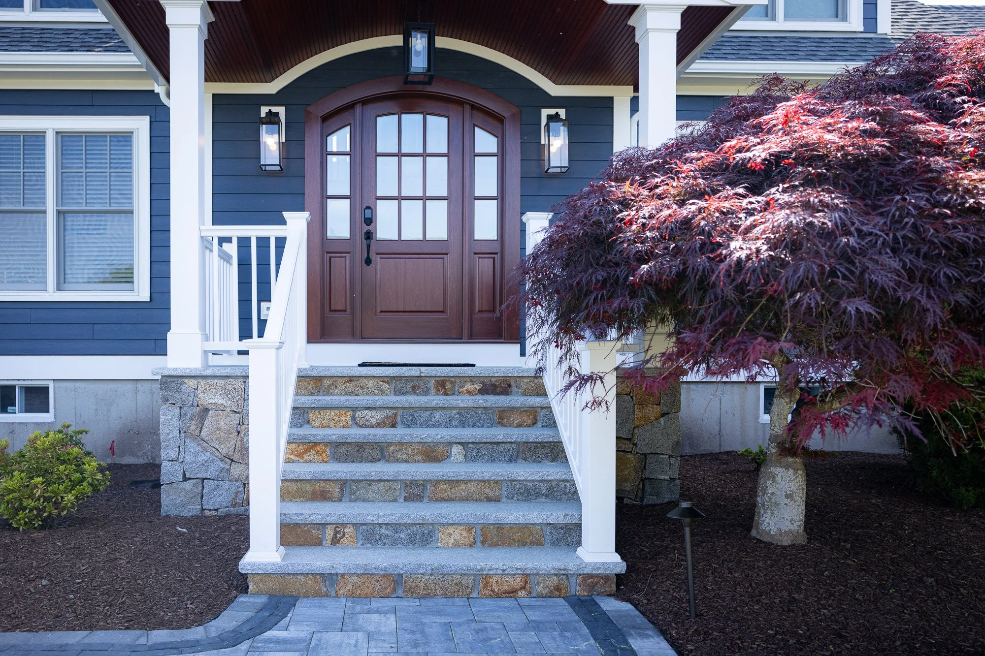 Front porch of a house with stone steps, white railings, a large red-leafed tree on the right, a dark blue house with white trim, and a wooden front door with glass panels, flanked by two windows and outdoor lighting fixtures.