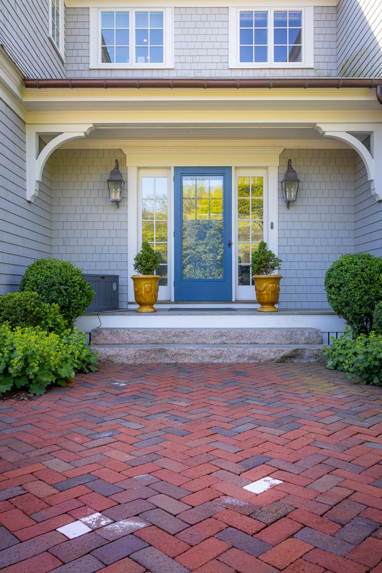 Front entrance of a house with a blue door, two potted plants, two wall lanterns, and a brick walkway.
