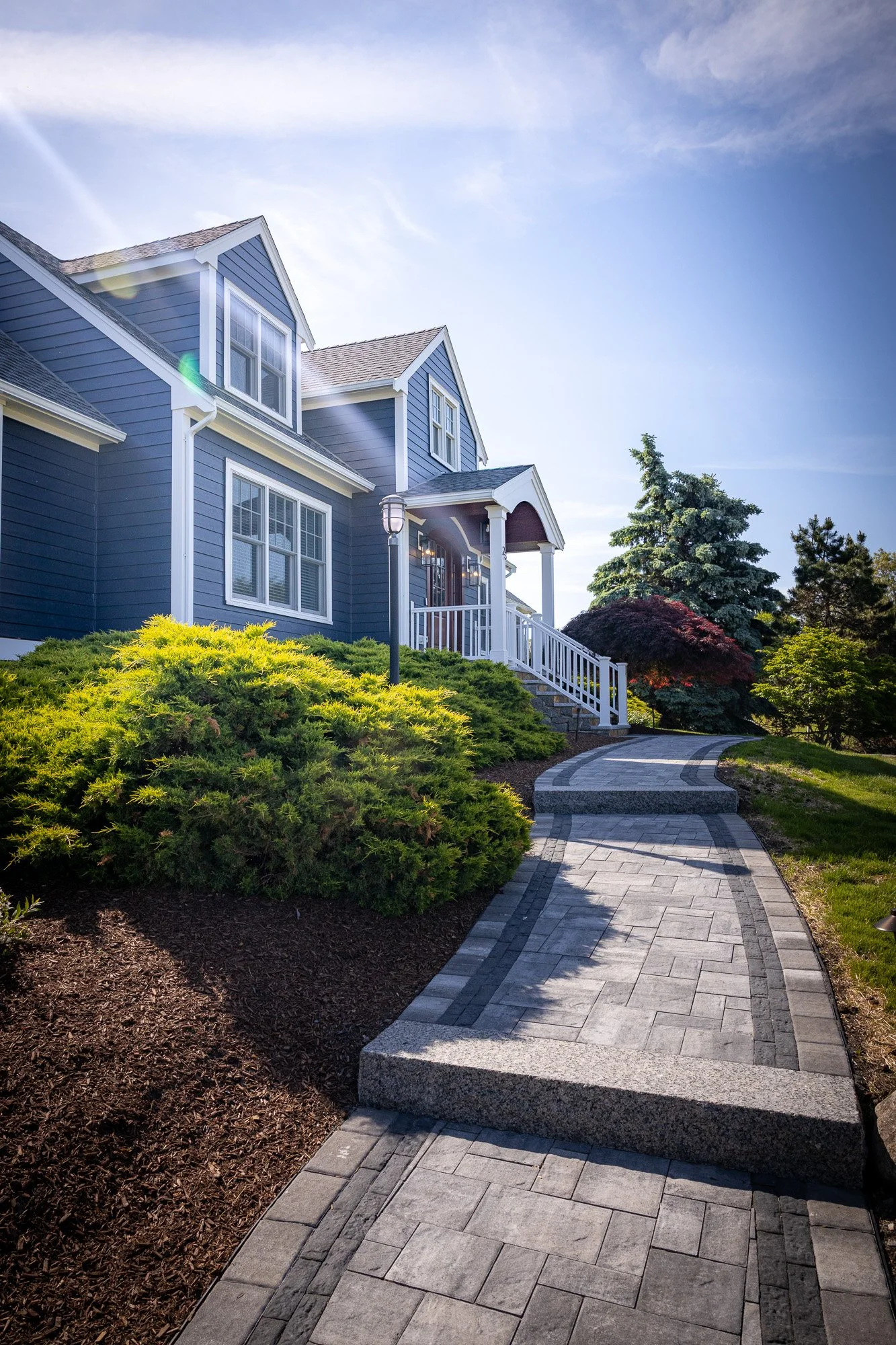 The image shows a modern blue house with white trim and multiple windows, surrounded by well-manicured green bushes and trees. A curved stone pathway leads to the front entrance, which has a staircase with white railing. The sky is clear with some wi