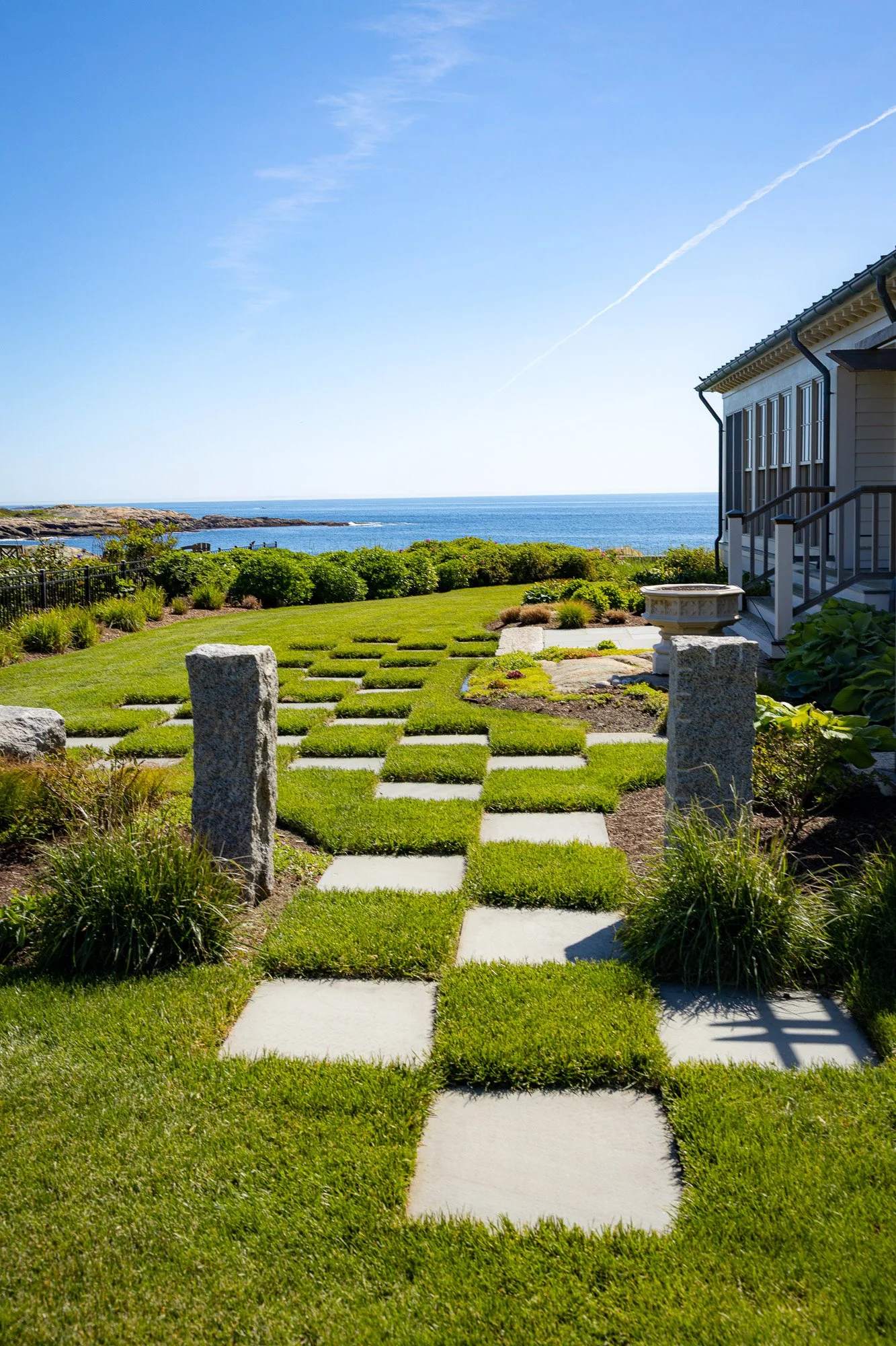 A beachfront garden view with a grassy lawn, stone pathway, and ocean in the background under a clear blue sky.