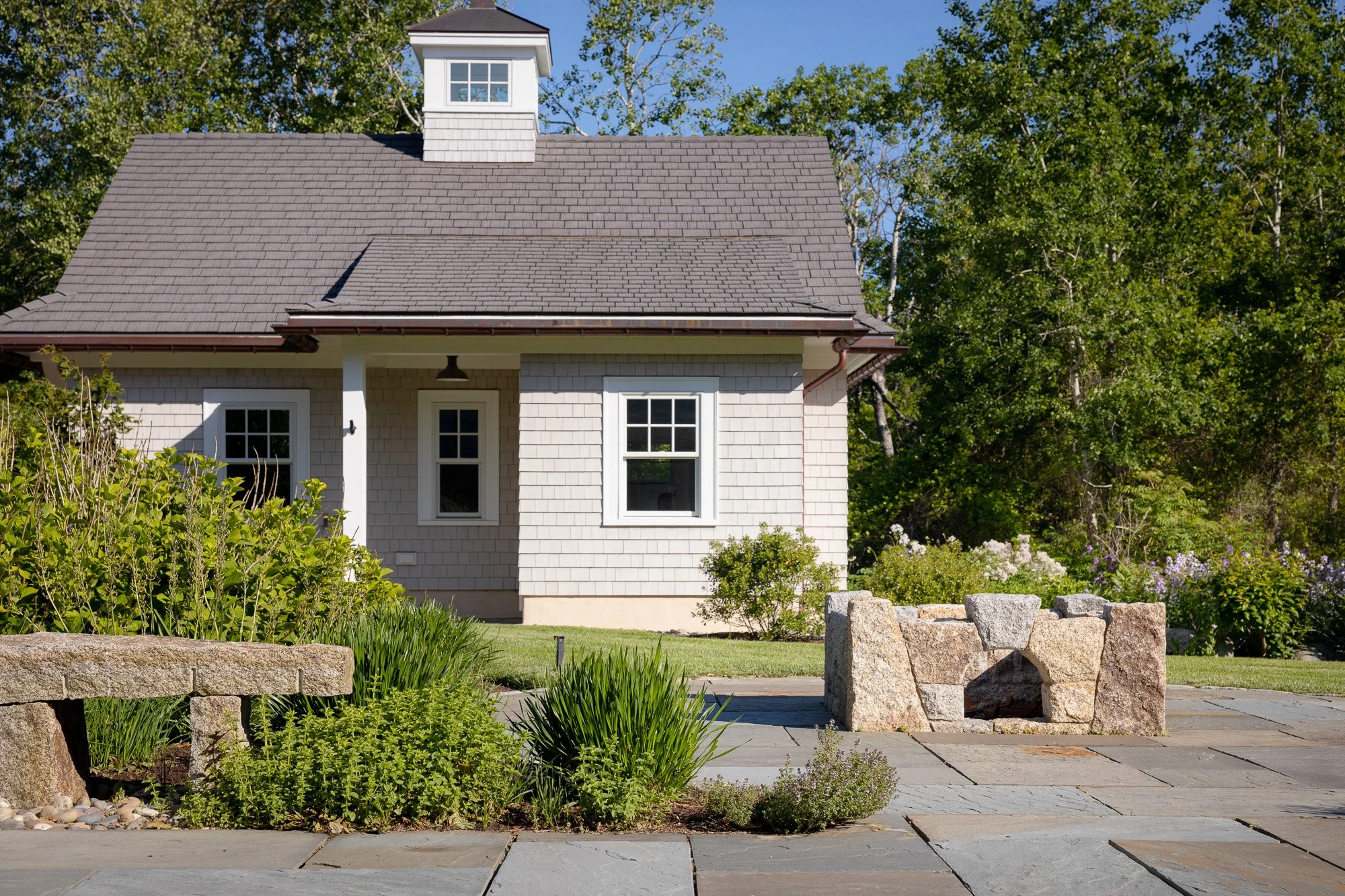 A house with a gray shingled roof, a chimney, white window frames, and a front porch, surrounded by a garden with green plants, a stone border, and a paved walkway, set against a background of green trees and a clear blue sky.