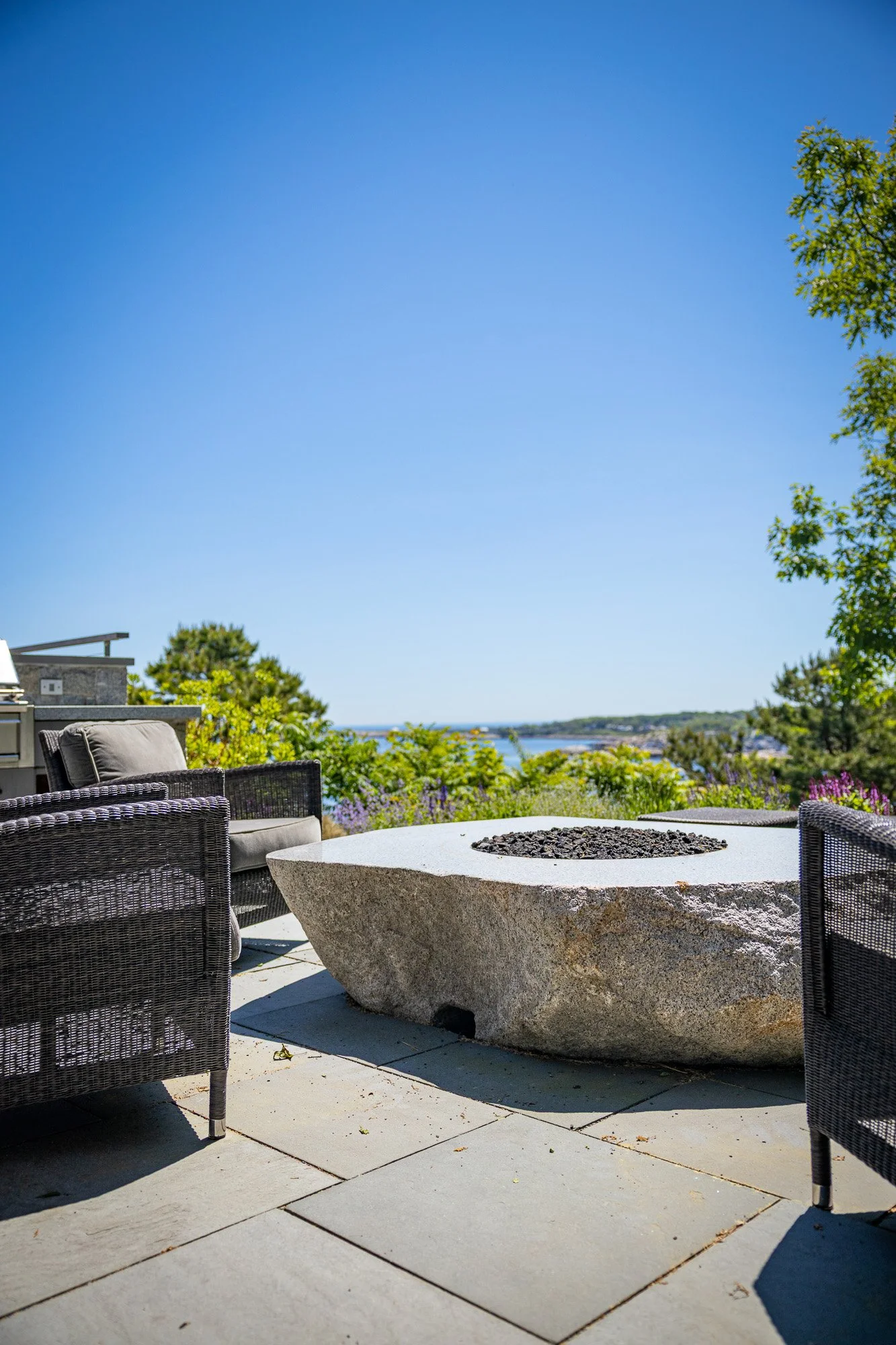 Outdoor patio area with woven chairs, a large stone fire pit, lush green trees, colorful flowers, and a view of the water and sky in the background.