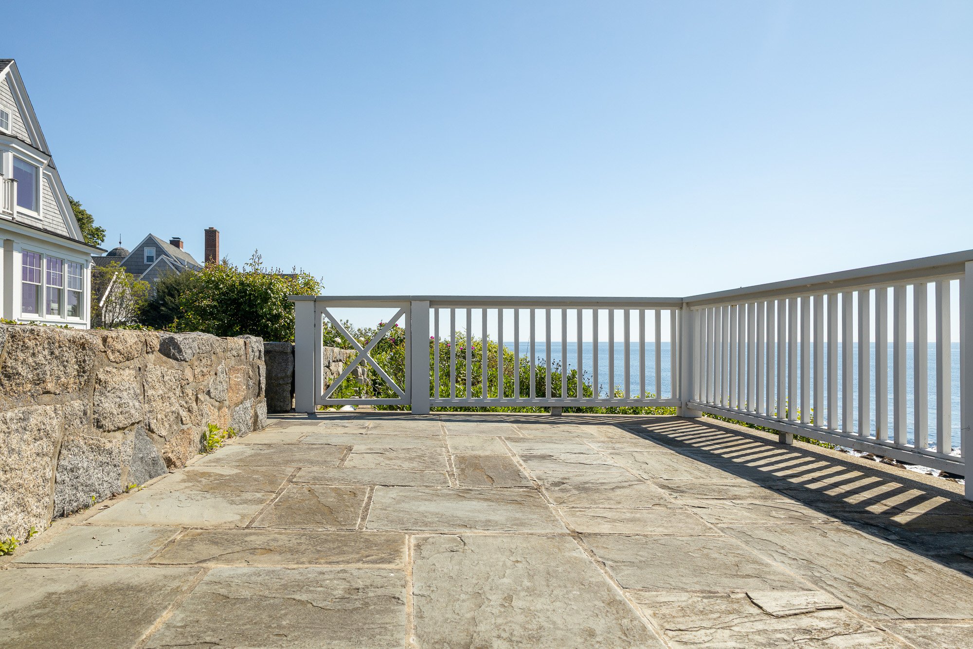 A stone patio with a white wooden railing overlooking the ocean, with houses and greenery visible in the background on a clear sunny day.