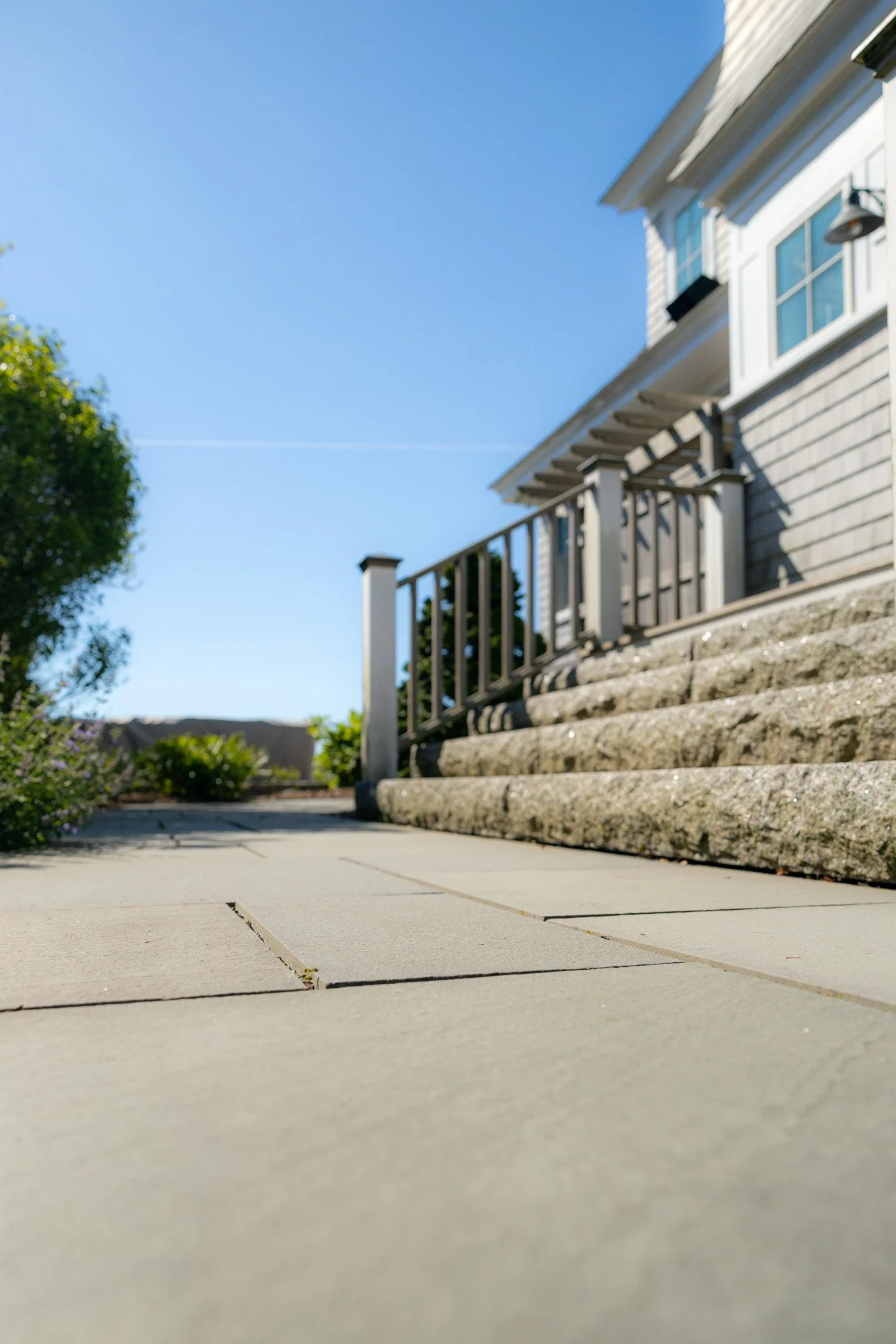 A house with a front porch, a staircase with stone steps, a wooden railing, and a white exterior wall with windows under a clear blue sky.