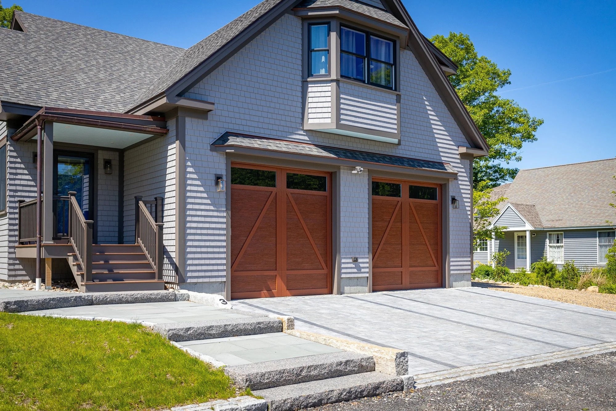 Front of a modern house with gray shingle siding, large brown garage doors, a small staircase leading up to a side door, and a driveway with a stone border. A small lawn in the foreground and neighboring houses in the background. Bright sunny day wit