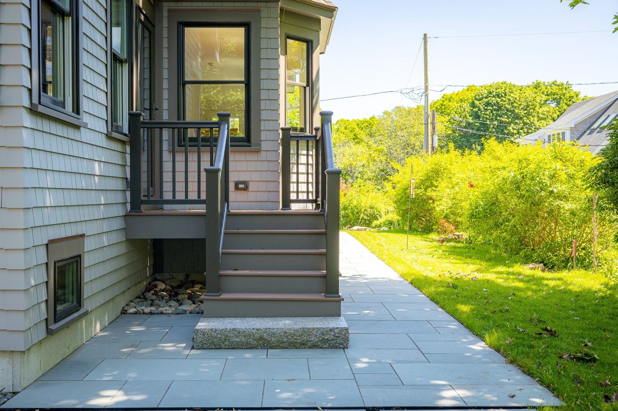 Backyard patio with stairs leading to a deck on a house with light-colored siding, green trees, and a grassy yard with a flagpole.