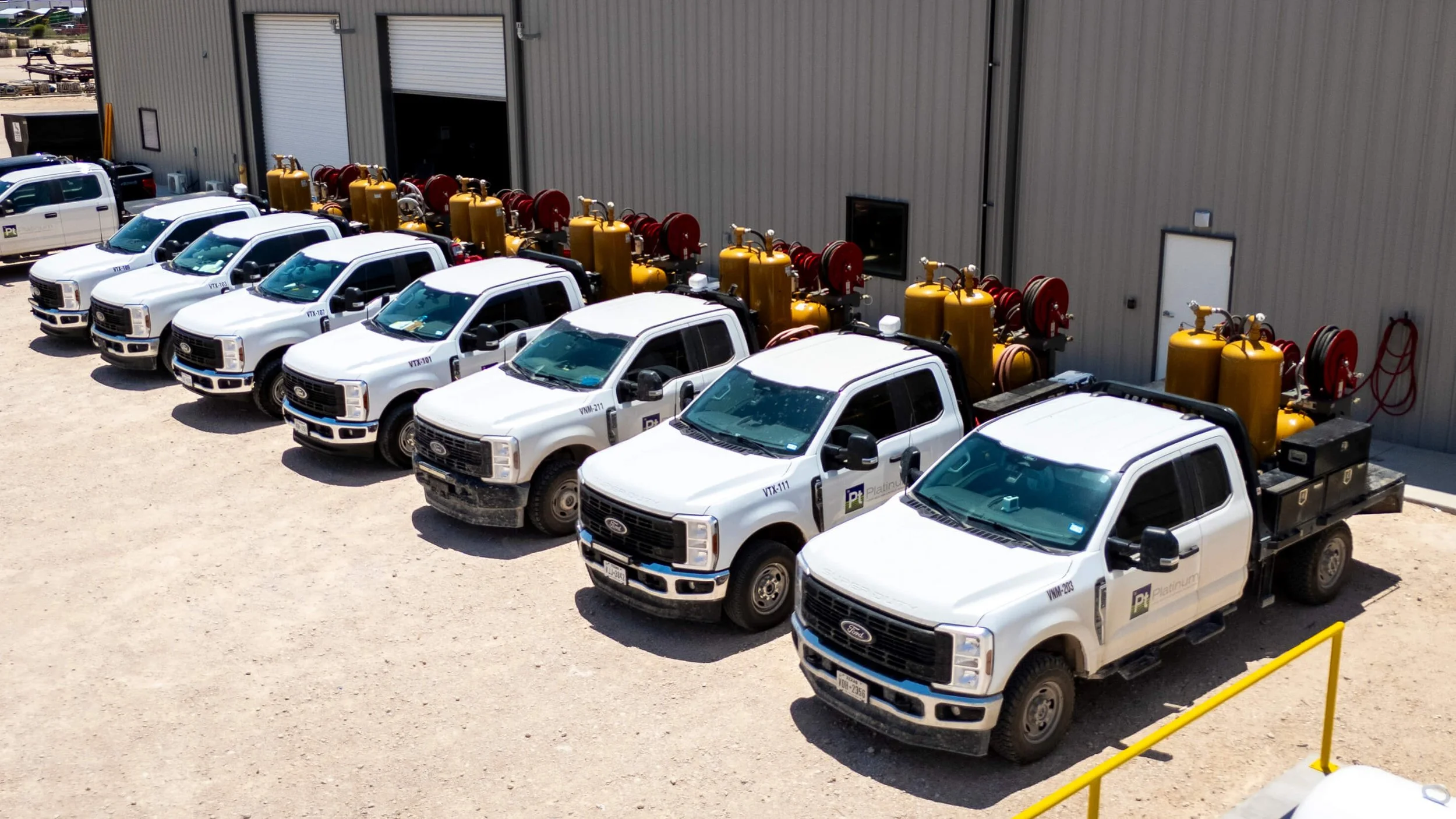 A row of white pickup trucks parked outside a large industrial building, with firefighter equipment and yellow tanks mounted on the trucks.