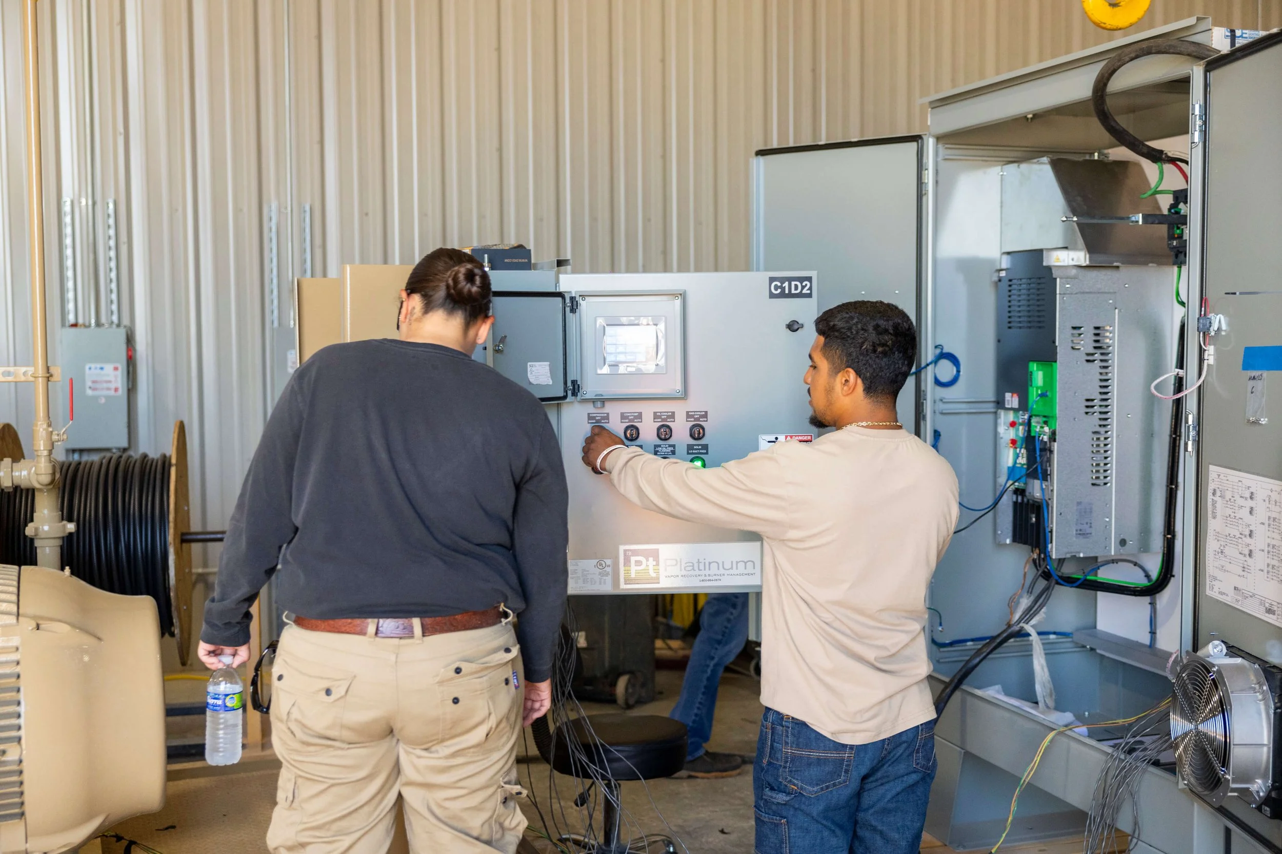 Two men working on electrical or mechanical equipment in an industrial setting, with a coach sitting nearby and water bottle on the floor.