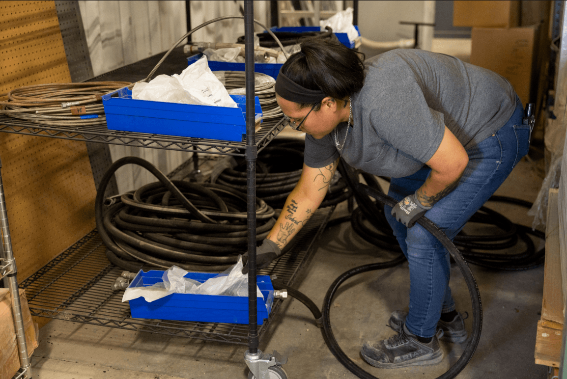 A woman with tattoos, glasses, and a headband is bending down to handle thick black hoses on a shelf in a warehouse or industrial setting.
