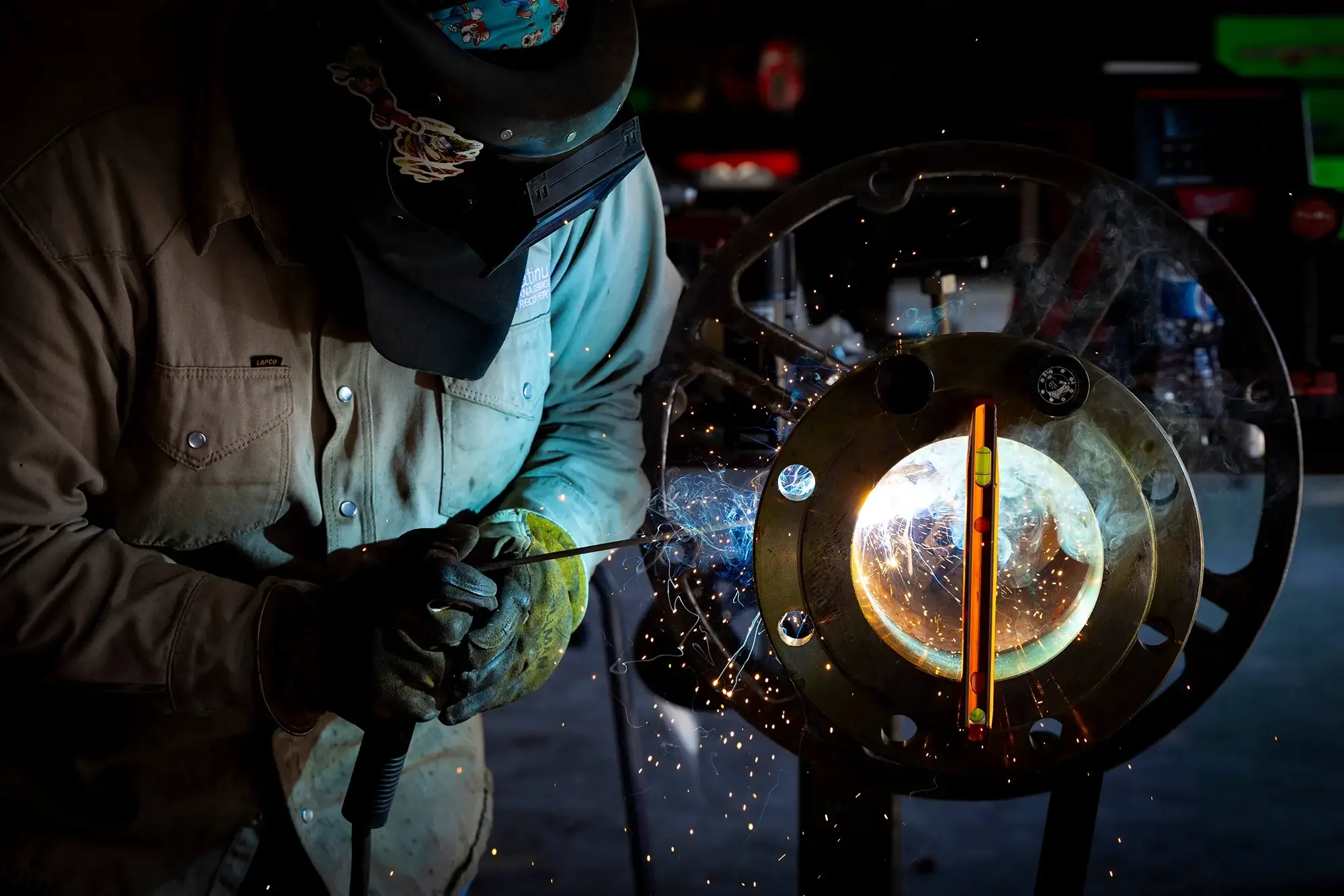 A worker welding a metal pipe, with sparks flying in the dark workshop.