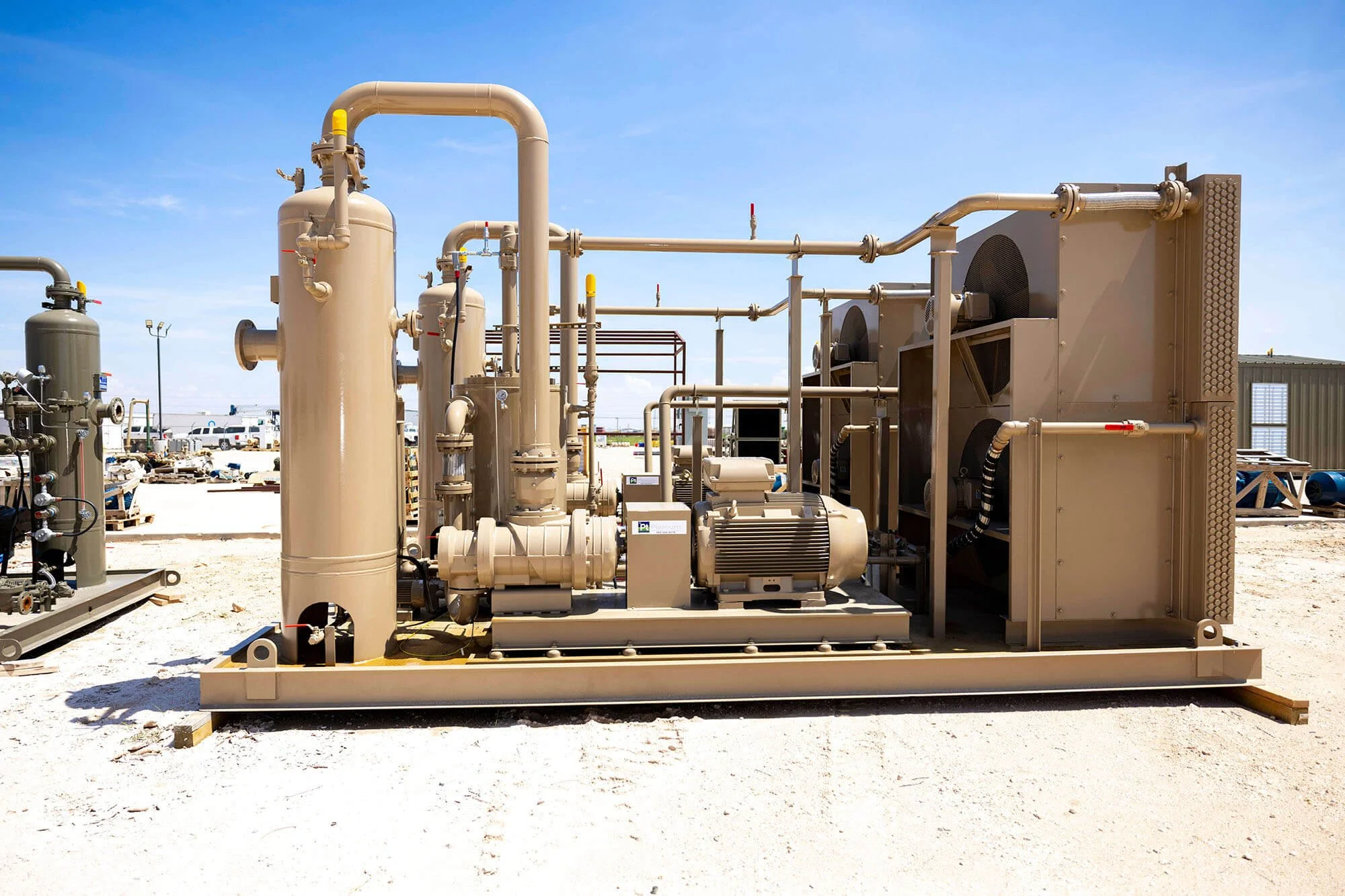 Industrial equipment with pipes, valves, and machinery outdoors on a sunny day, with a building and parked cars in the background.