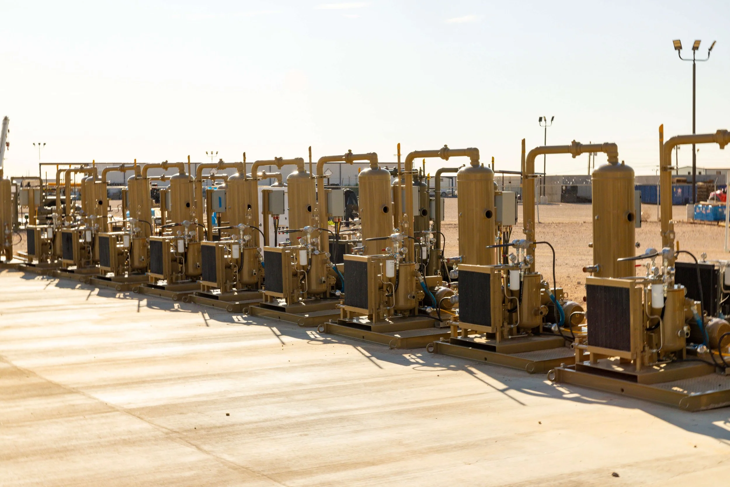 A row of industrial equipment with yellow pipes and control boxes installed on a concrete surface outdoors, with a wire fence and various structures in the background under a clear sky.