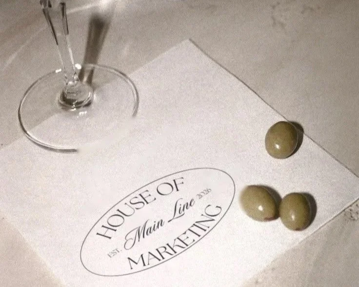 A wine glass with cocktail picks, a piece of paper with 'House of Main Lime Marketing' printed on it, and three olives placed on the paper.