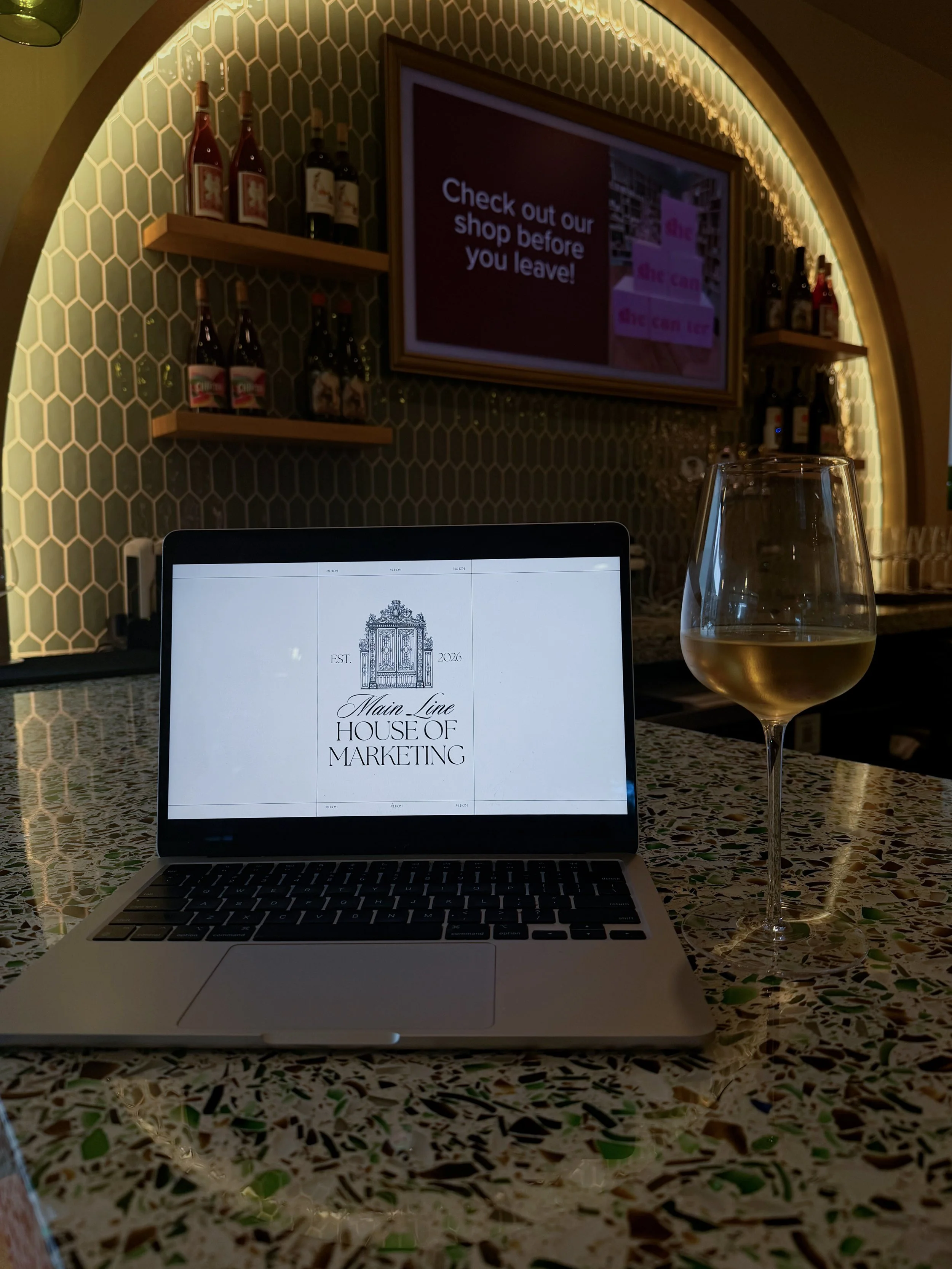 A laptop displaying the logo and text for Main Line House of Marketing, a glass of white wine, and a blurred background with shelves of bottles and a digital screen.