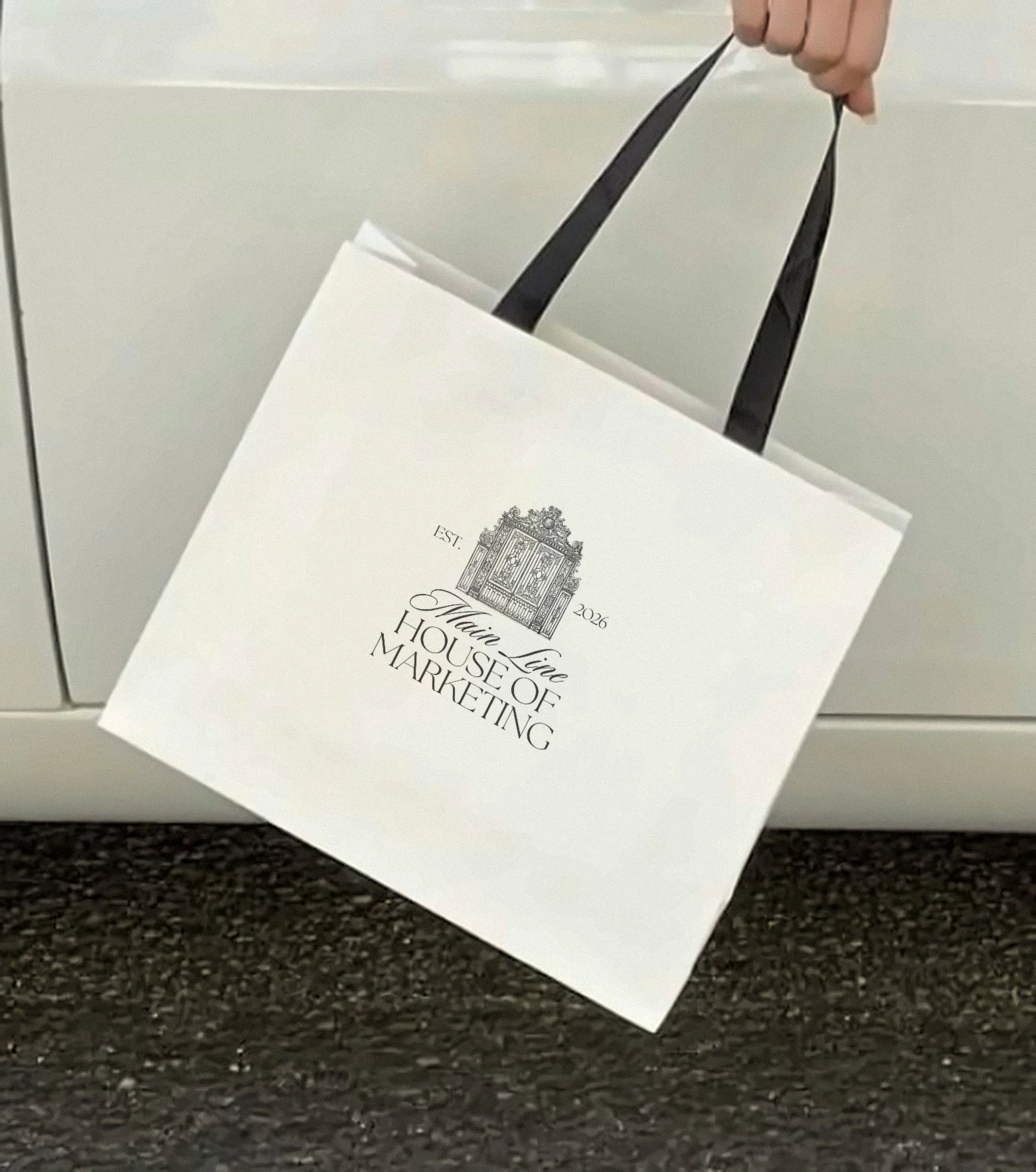 A white paper bag with black handles hanging out of a white mailbox. The bag has text and a decorative illustration, which reads "Est. 2026 Main Line"House of Marketing".