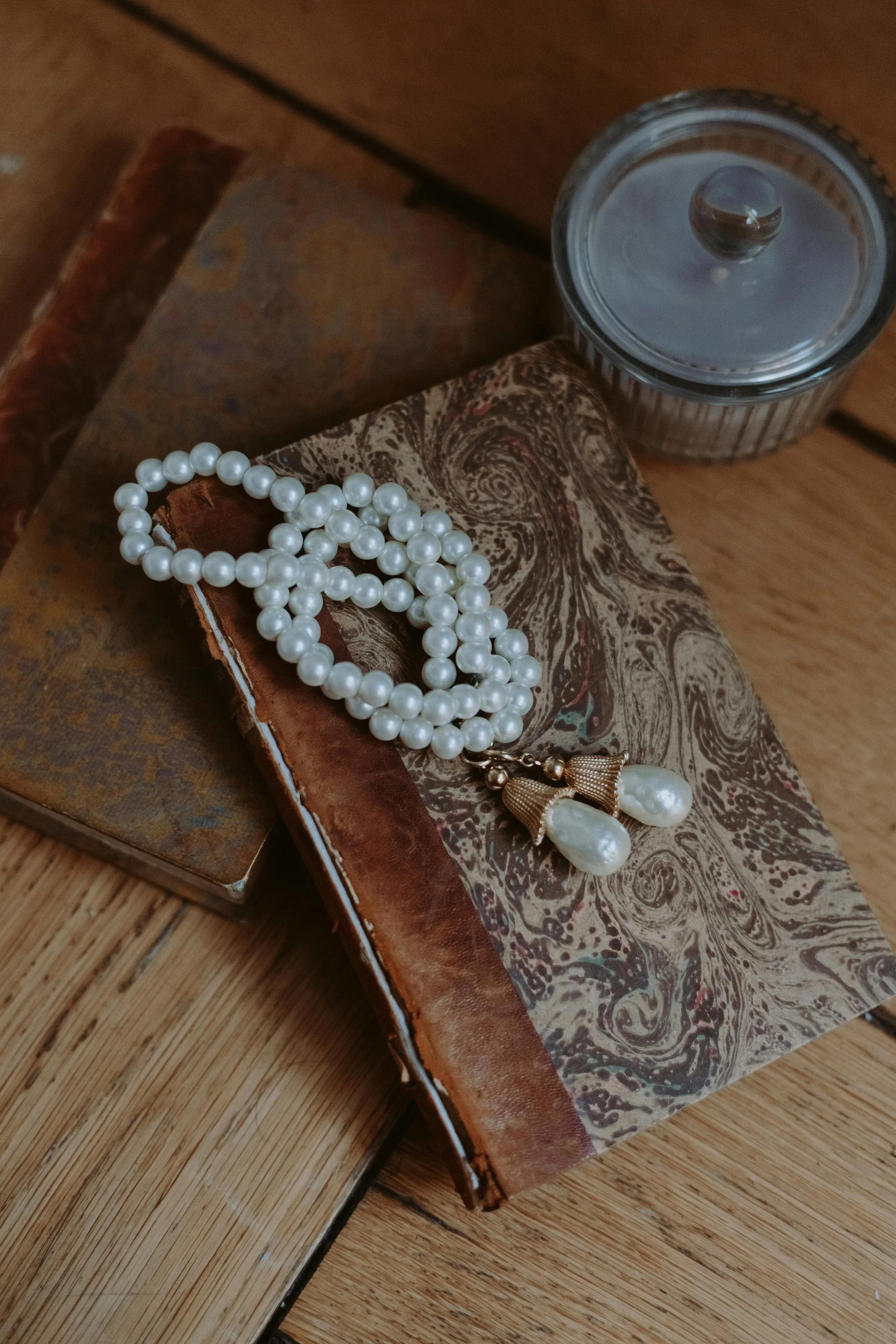 A string of white pearls with a golden tassel, a small vintage book with a marbled cover, and a clear glass container with a black lid are arranged on a wooden surface.