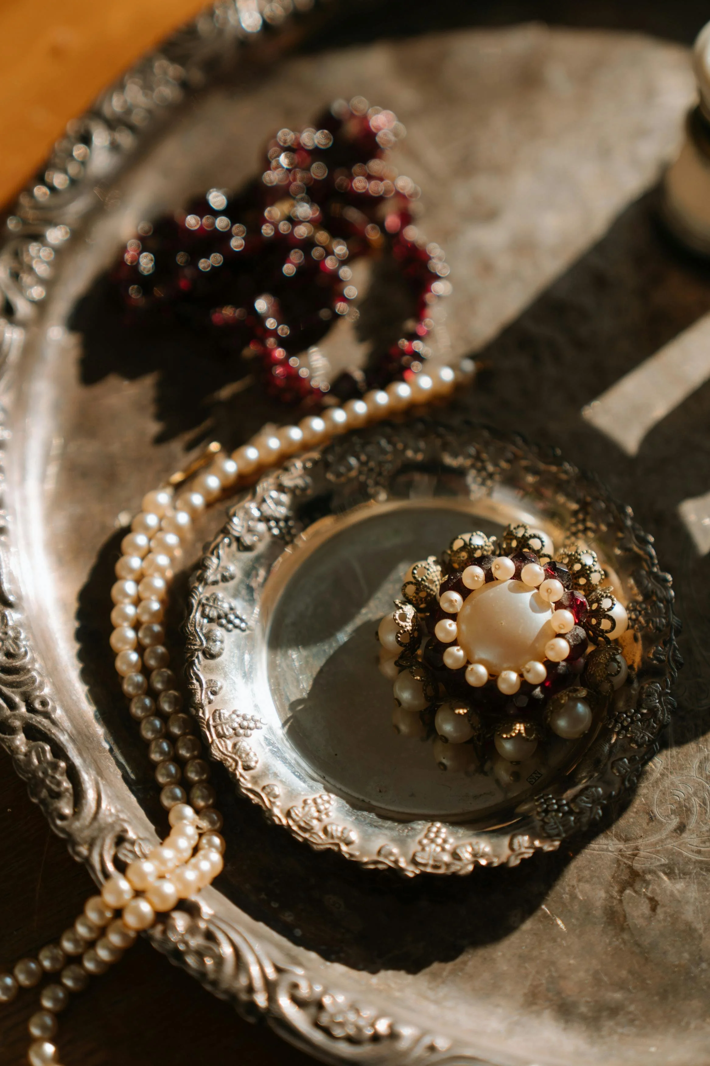 Close-up of jewelry, including pearl necklaces and a brooch with pearls and red stones, on an ornate silver tray.
