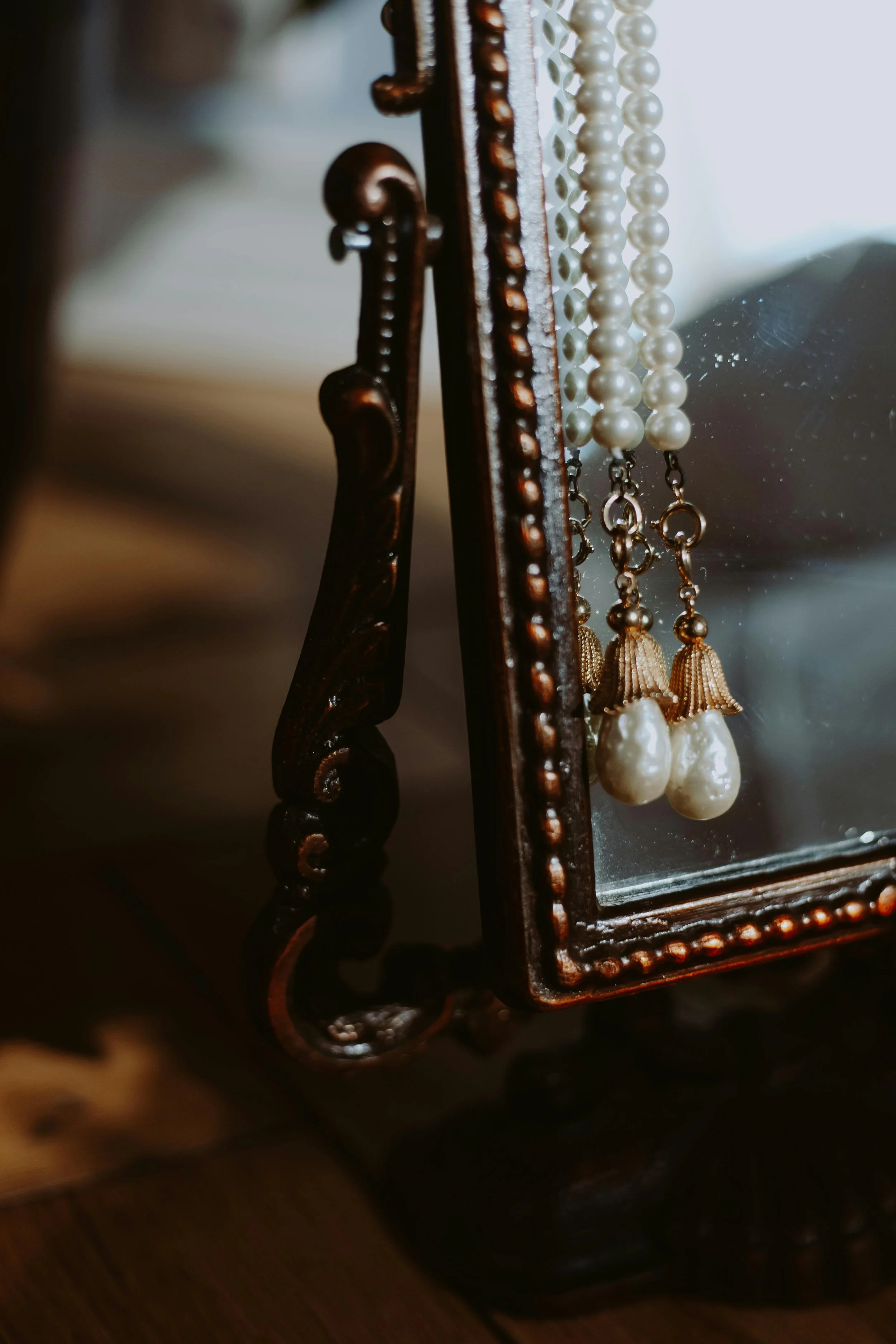 Close-up of jewelry hanging on a mirror frame, including pearl necklaces and a pair of pearl earrings, with a decorative, dark wooden frame.