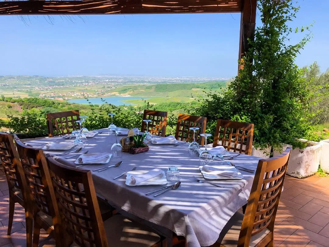 Outdoor dining table set with white tablecloth, napkins, glasses, and utensils on a terrace overlooking a green landscape with a lake and distant town under a blue sky.