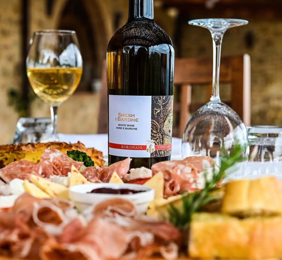 A bottle of white wine on a table surrounded by glasses, cheese, meats, and a bowl of berry jam at a rustic dining setting.