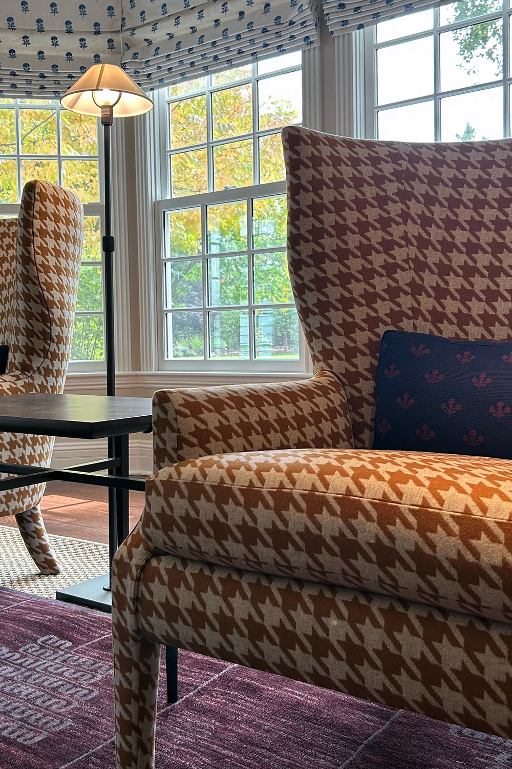 A cozy living room with armchairs featuring houndstooth patterns, a floor lamp, large windows with floral curtains, and a view of trees outside.