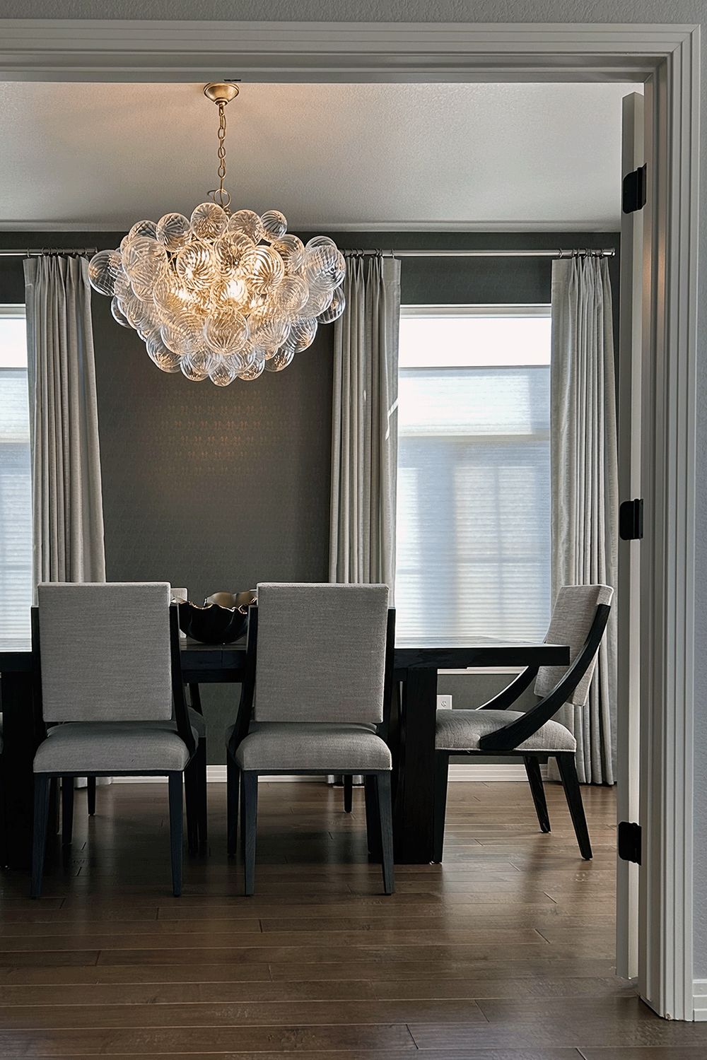 Dining room with a dark wood table, beige upholstered chairs, linen drapery, a modern chandelier, and hardwood floors.