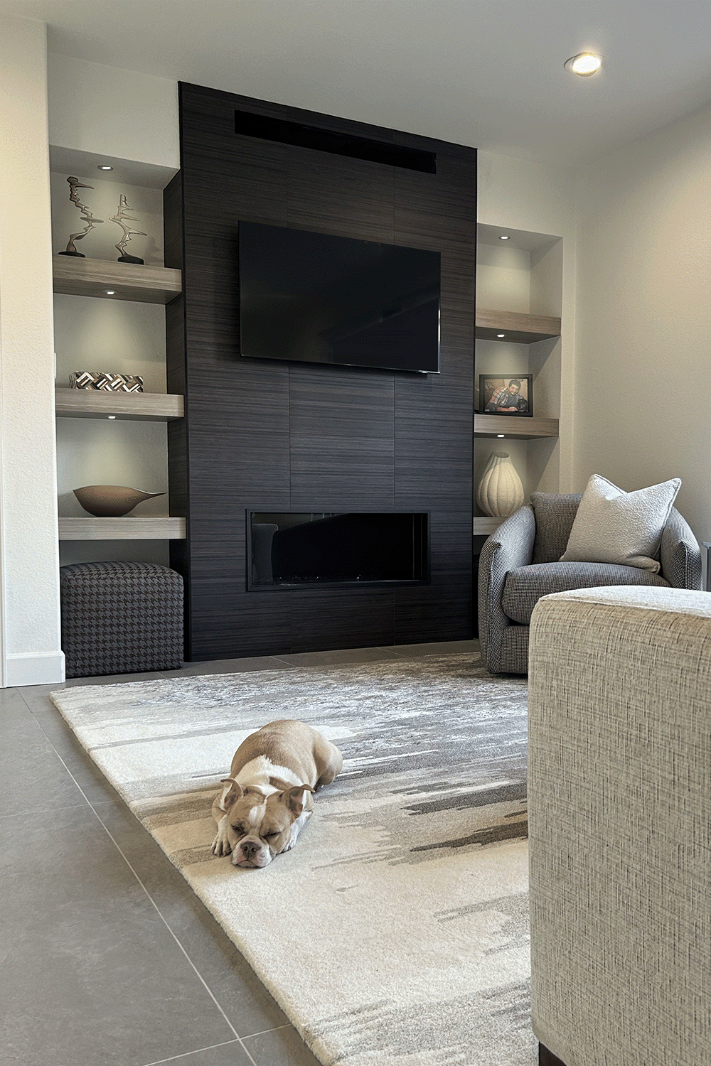 A living room with a tan and white bulldog sleeping on a patterned area rug, a gray armchair with pillows, a beige couch, built-in shelves with decorative objects, artwork, a cabinet, a black tiled fireplace, and a mounted flat-screen TV on a dark wo