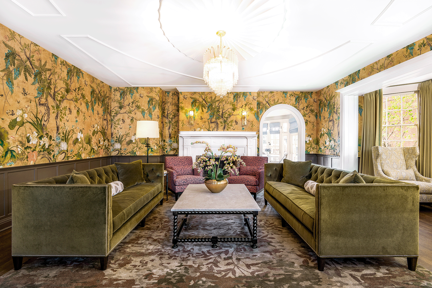 Green velvet tufted sofas frame an ornately milled fireplace and oval glass chandelier.